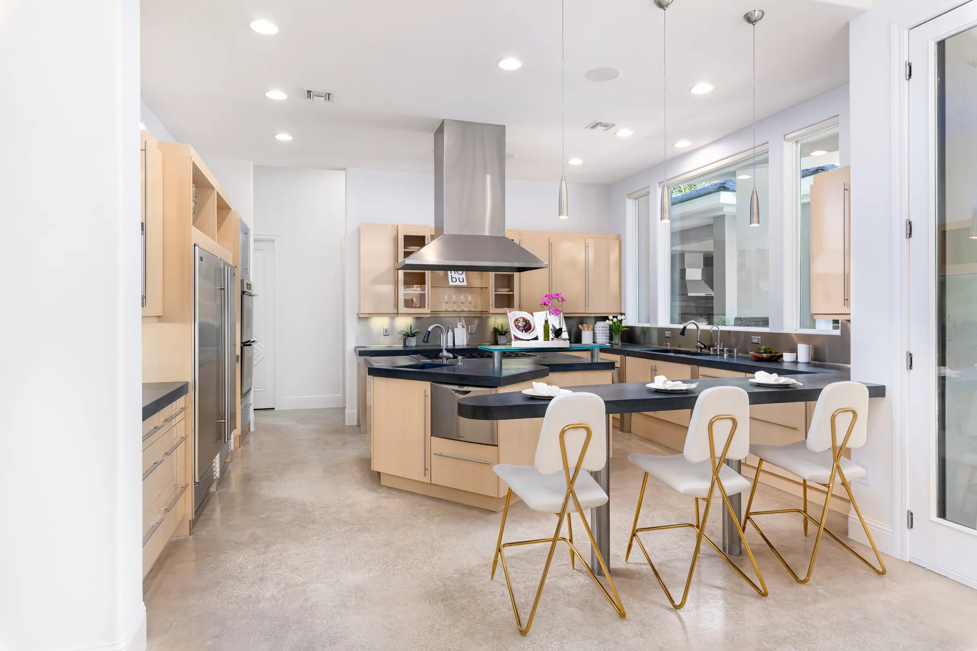 A spacious modern kitchen with light wood cabinetry, black countertops, stainless steel appliances, and a large island with white and gold barstools.
