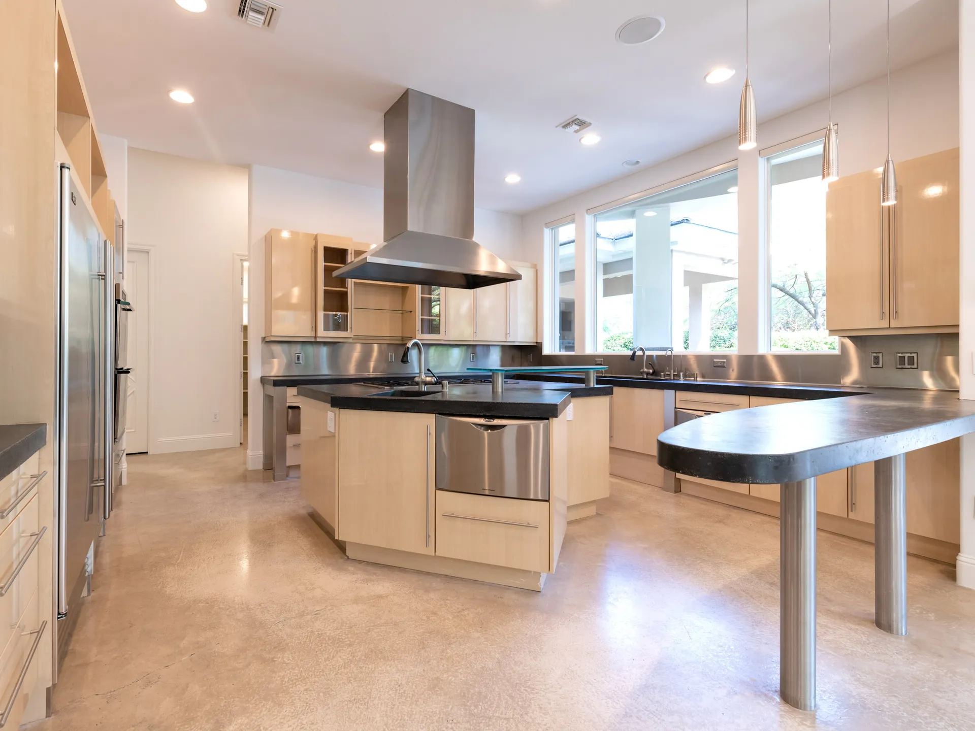 An open kitchen with light wood cabinetry, black countertops, stainless steel appliances, and large windows allowing in natural light.
