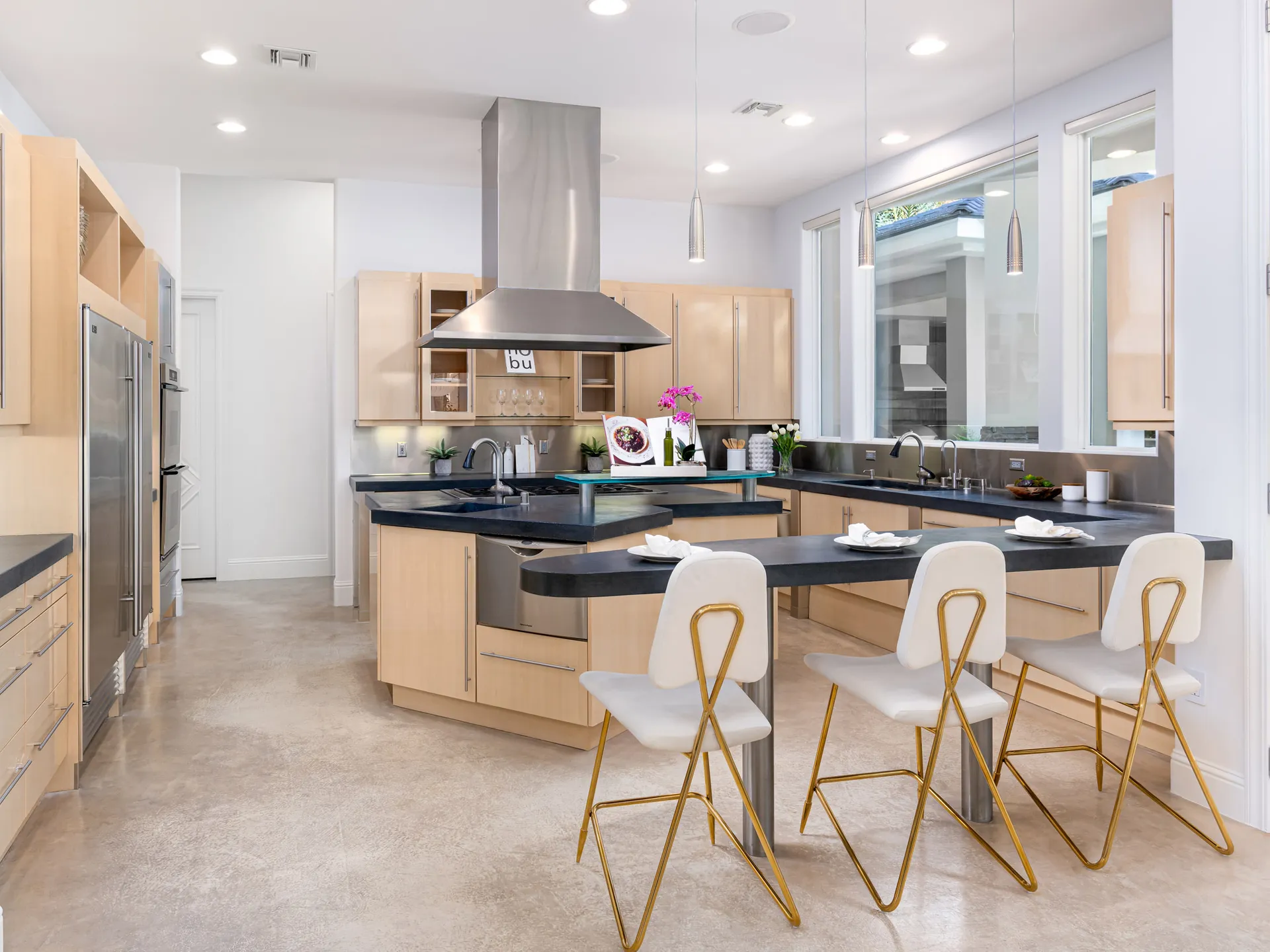 A spacious kitchen with light wood cabinetry, black countertops, a large island with seating, and stainless steel appliances under sleek pendant lighting.