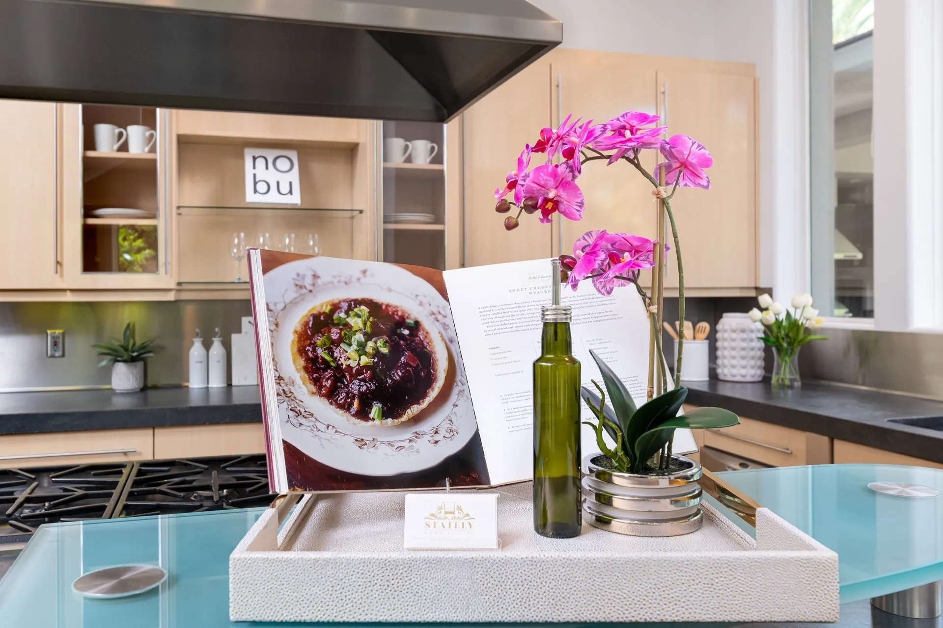 A modern kitchen display featuring an open cookbook, a glass bottle of olive oil, and a vibrant pink orchid on a decorative tray.