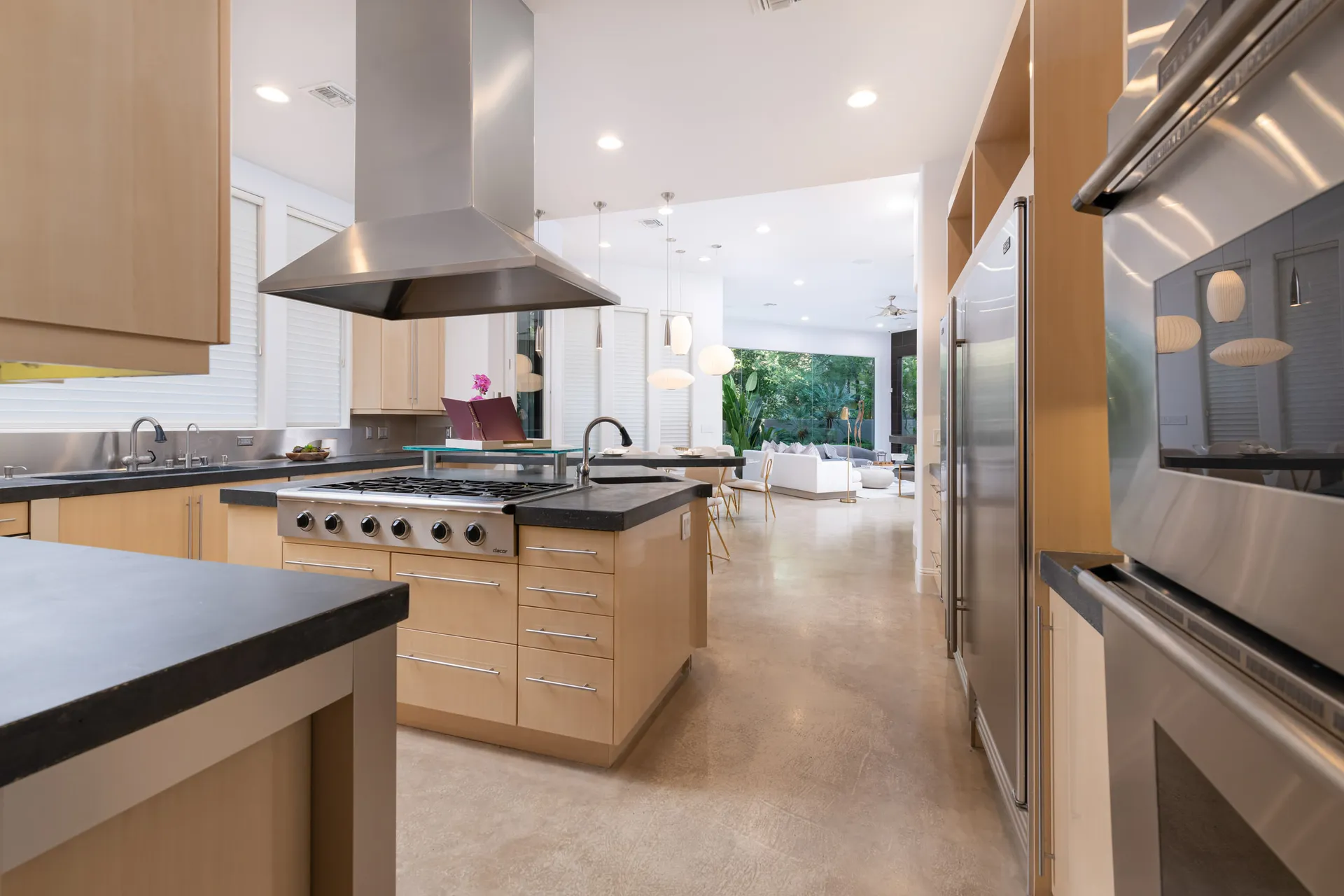 A contemporary kitchen with light wood cabinetry, black countertops, stainless steel appliances, and an open layout leading to a bright living area.