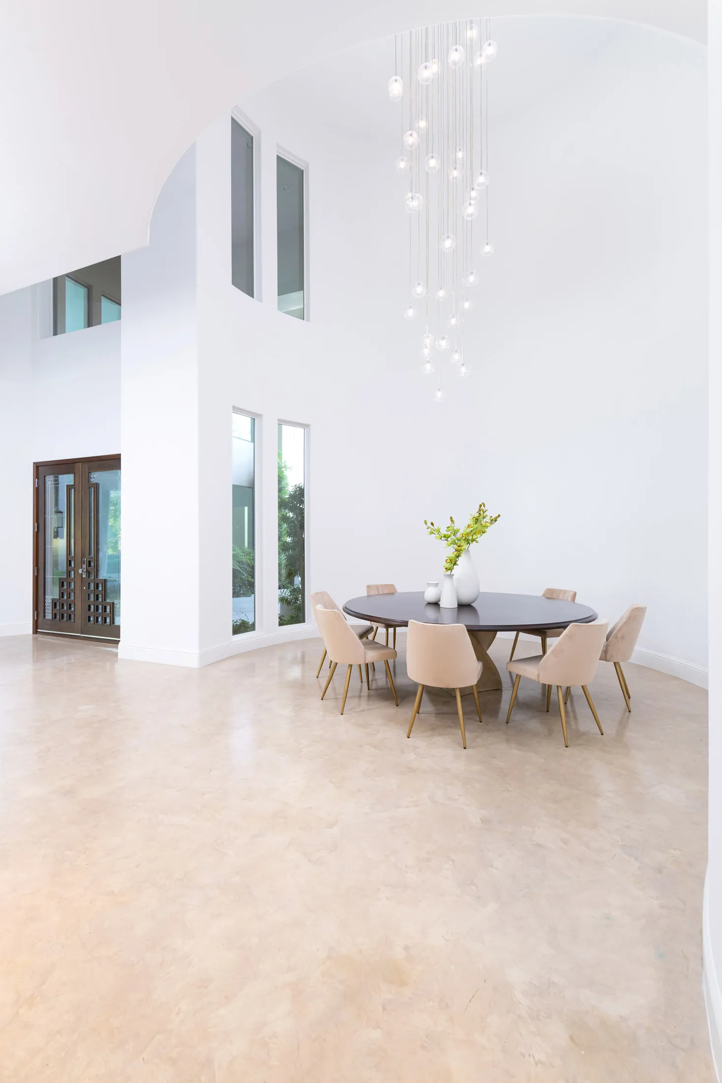 A spacious dining area with a round dark wood table, beige upholstered chairs, and a cascading glass pendant light against a high-ceiling white interior.