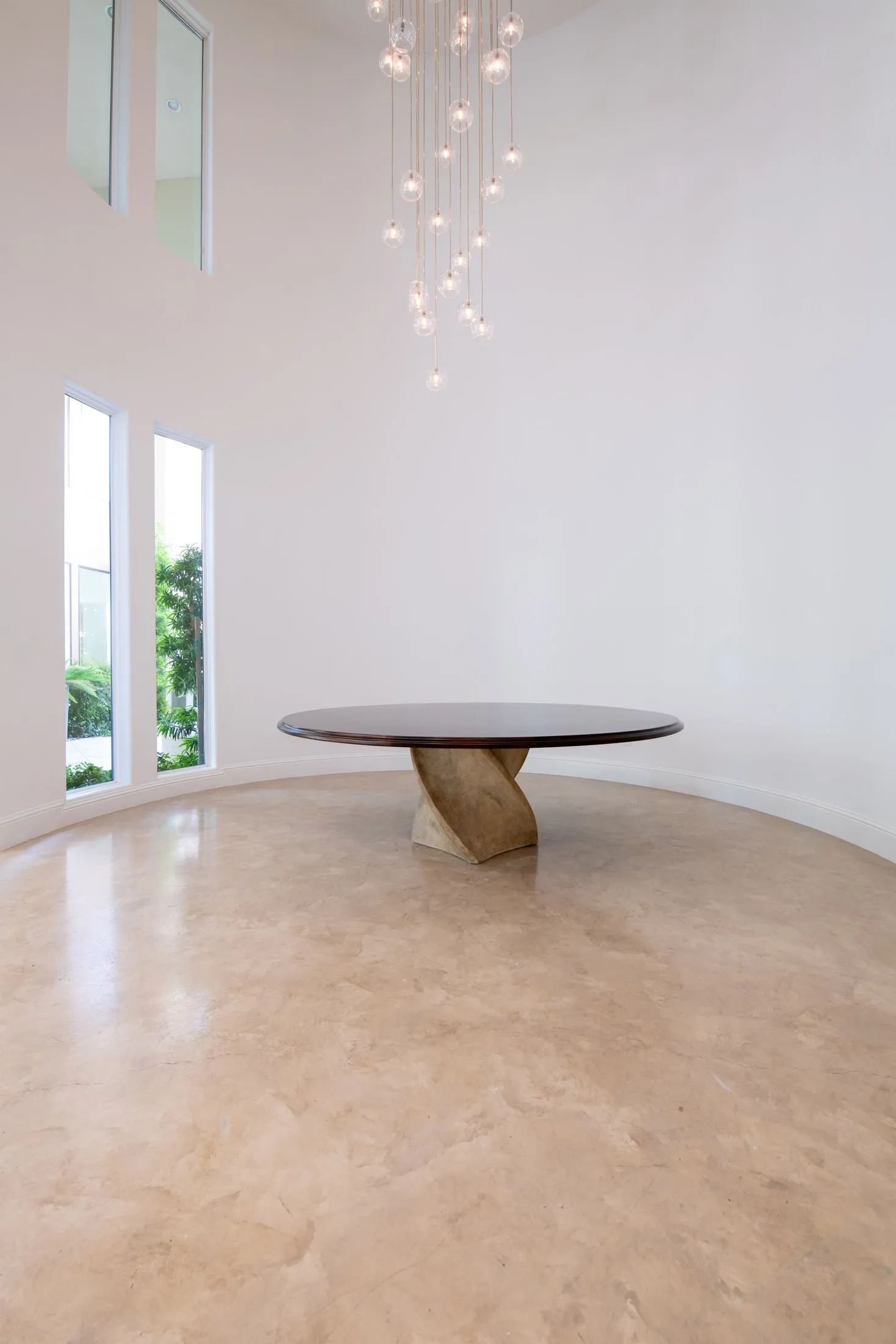 An empty dining room with a sculptural round table, high ceilings, cascading glass pendant lighting, and tall windows bringing in natural light.