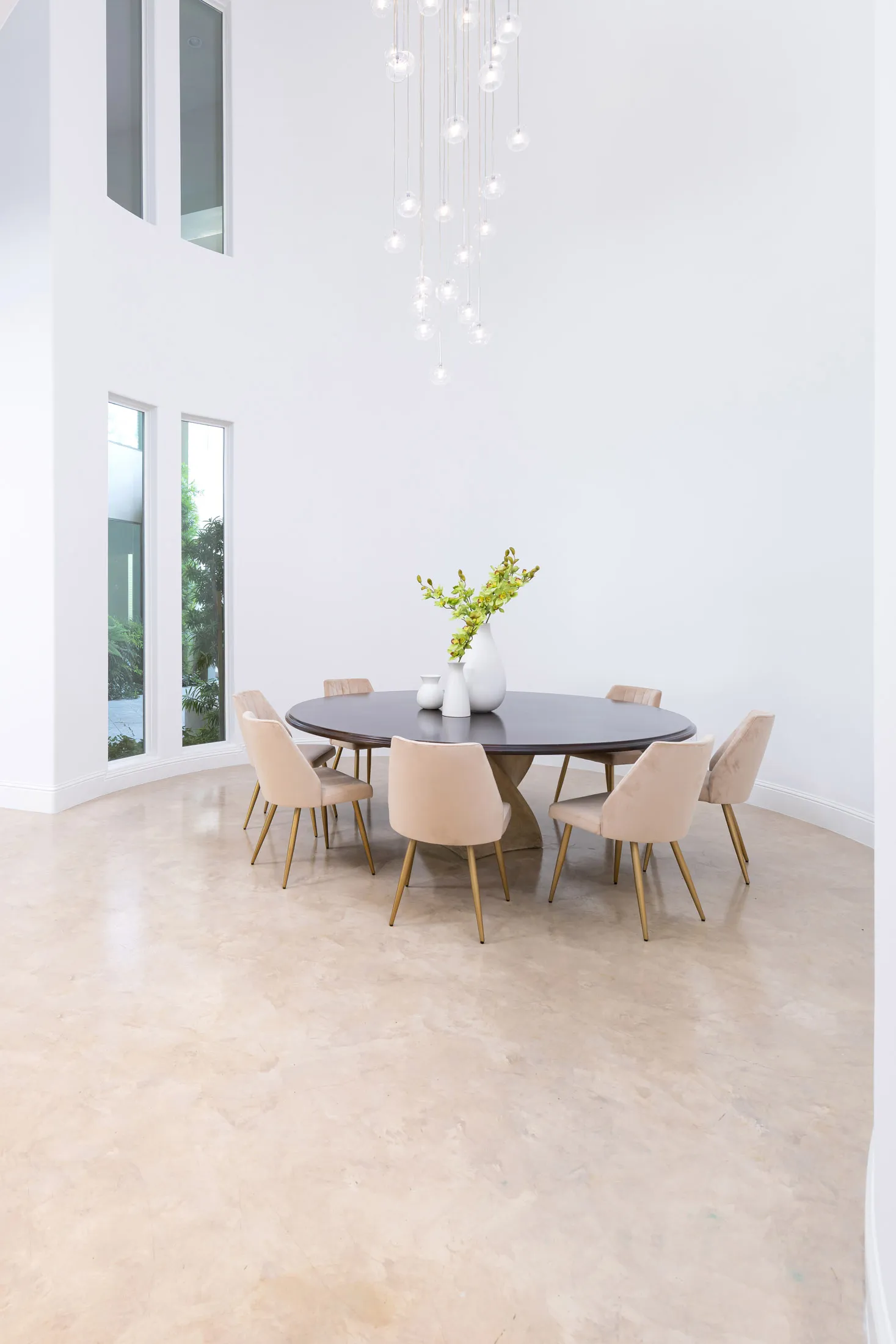 A contemporary dining space featuring a round dark wood table, beige velvet chairs with gold legs, and a cascading glass pendant light.