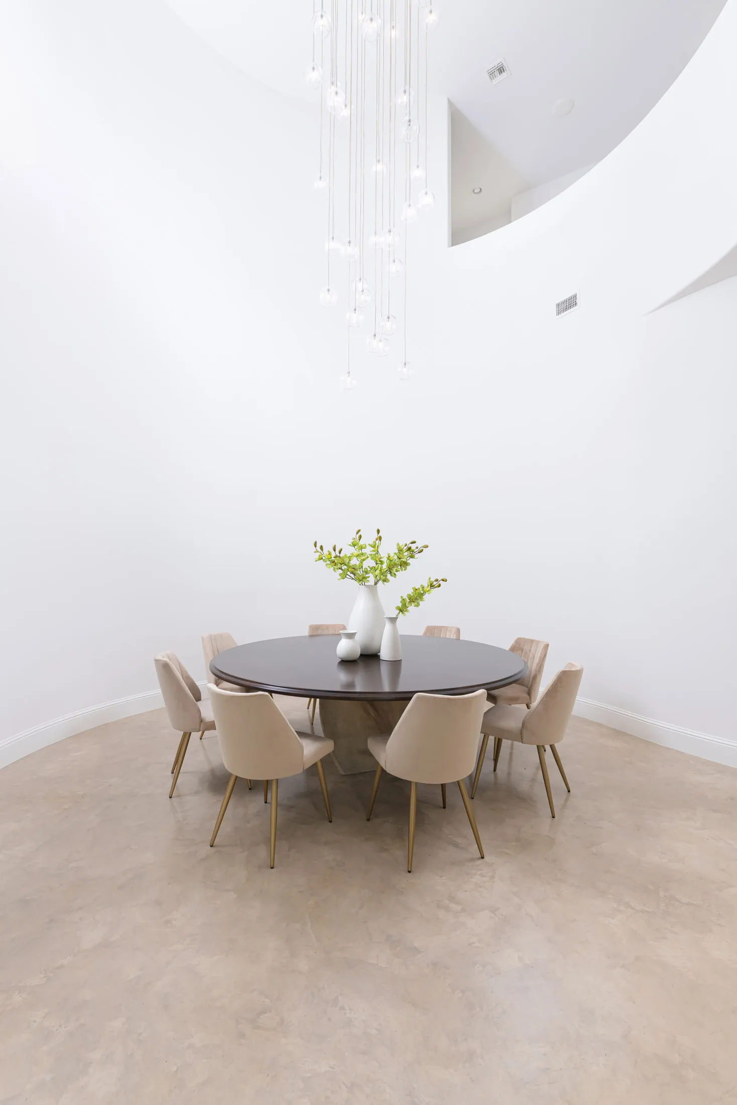 A contemporary dining area featuring a round dark wood table, plush beige chairs with gold legs, and a cascading glass pendant light.