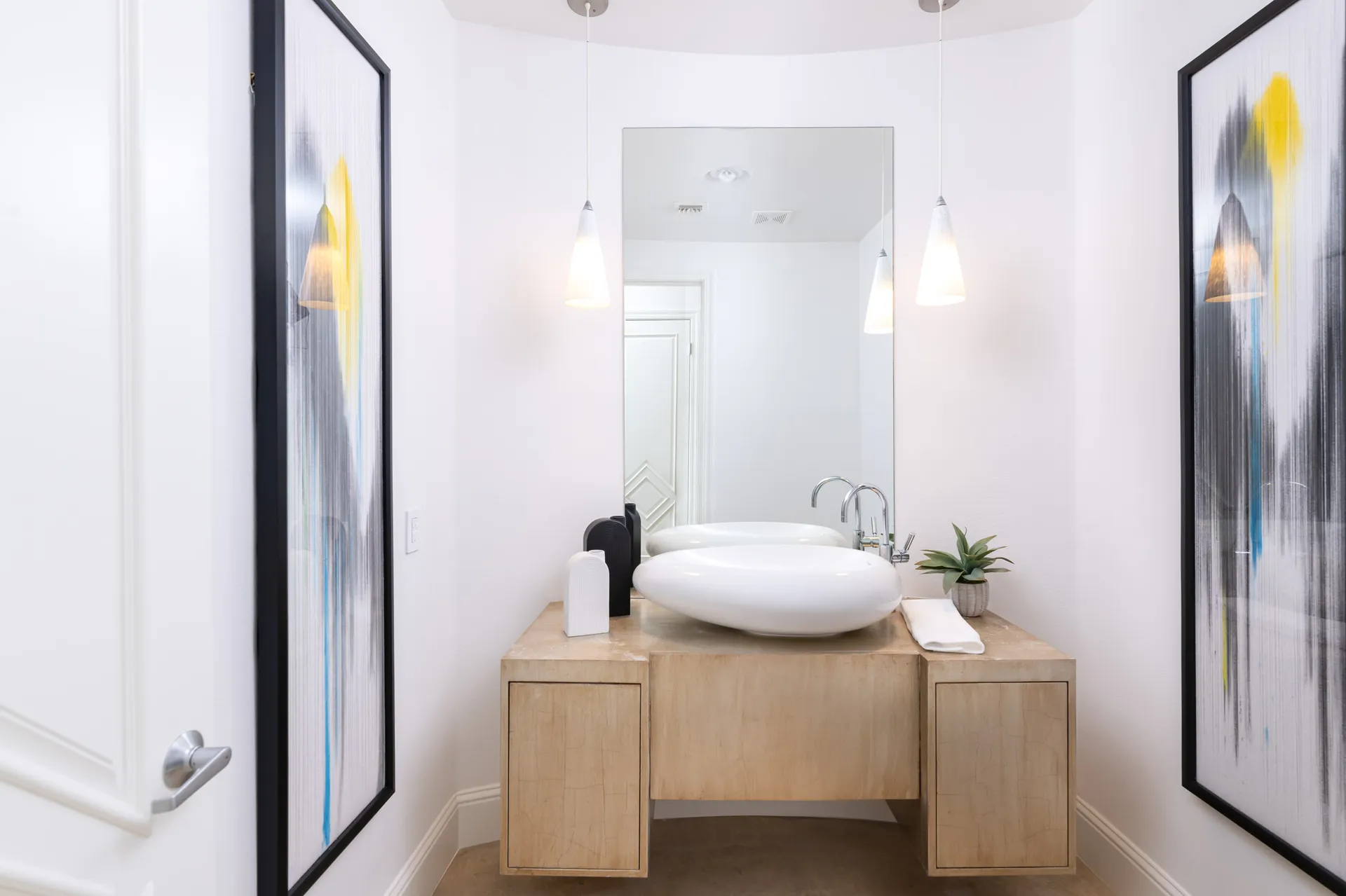A sleek bathroom featuring a floating wood vanity, an oval vessel sink, large mirror, and bold abstract artwork on both walls.