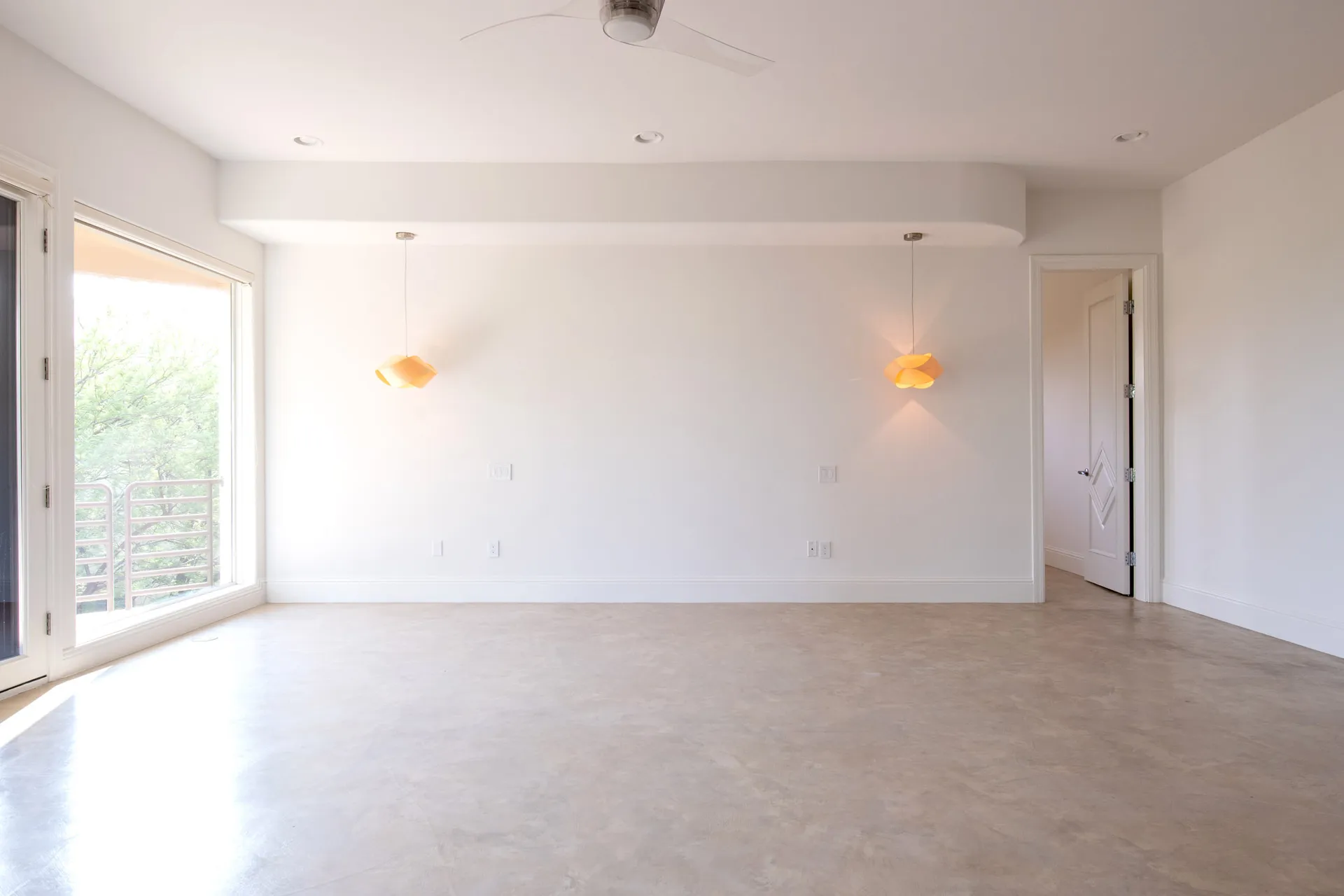 An empty master bedroom with white walls, a concrete floor, large windows, and two modern pendant lights.