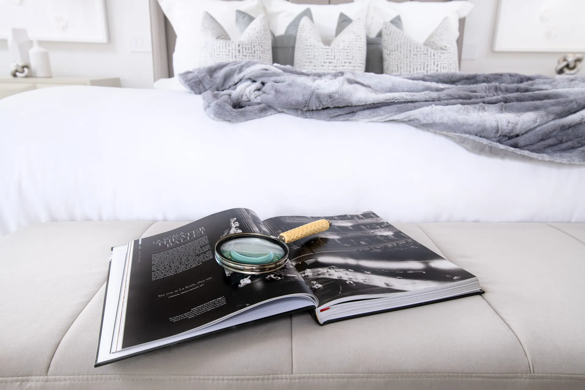 A neatly styled bedroom bench with an open book, a magnifying glass, and a plush bed with soft pillows and a textured throw blanket.