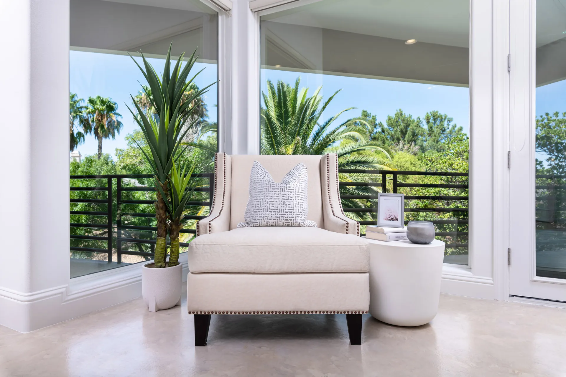 A cozy reading nook featuring a cream upholstered chair, a white side table with decor, and a potted plant, set against large windows with lush greenery.