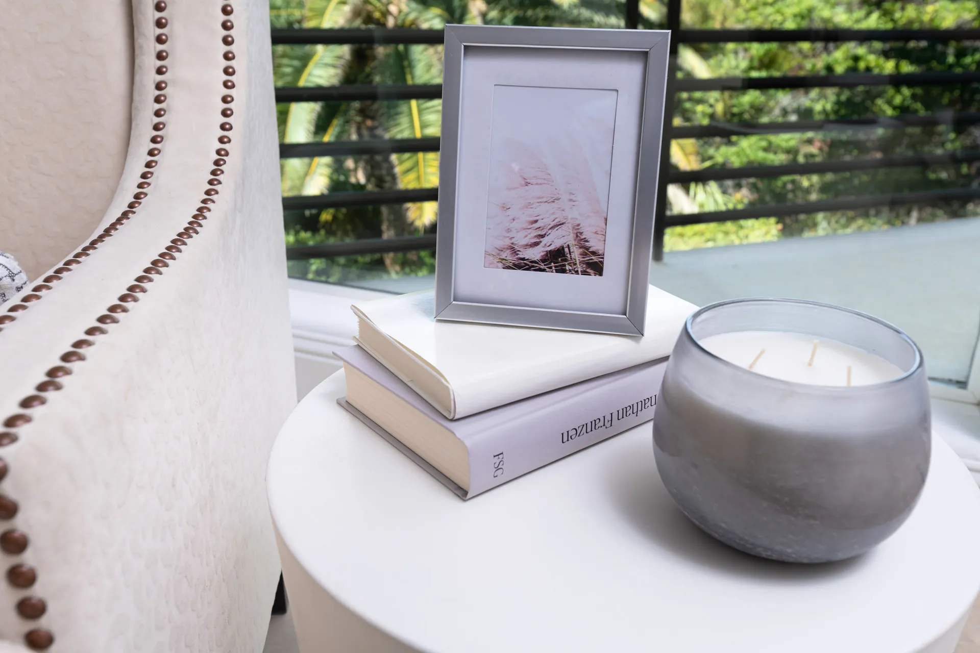 A white side table styled with stacked books, a silver picture frame, a glass candle, and a cream upholstered chair with nailhead trim.