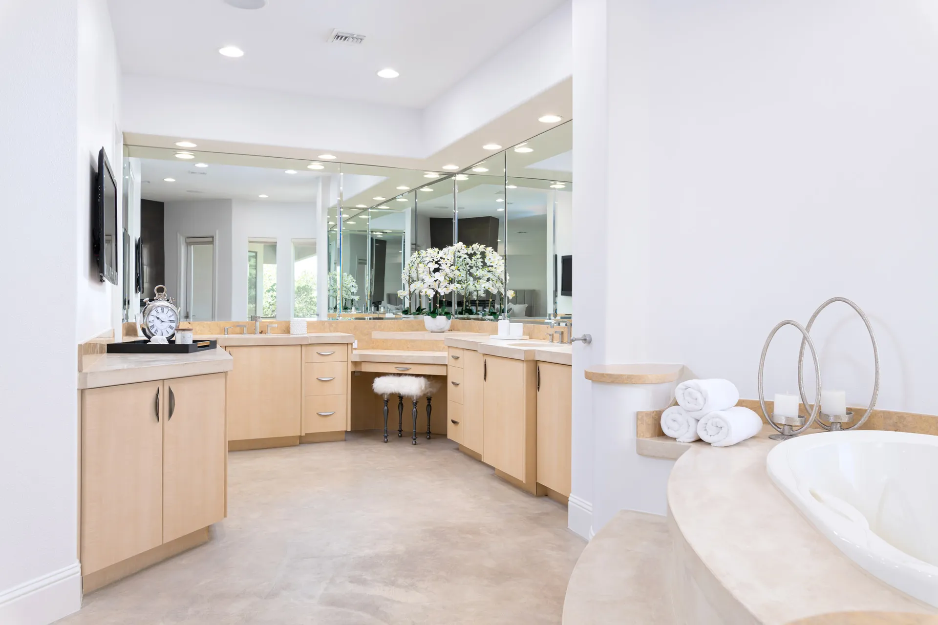 A spacious master bathroom featuring light wood cabinetry, a built-in bathtub, a mirrored vanity, and soft decorative accents.