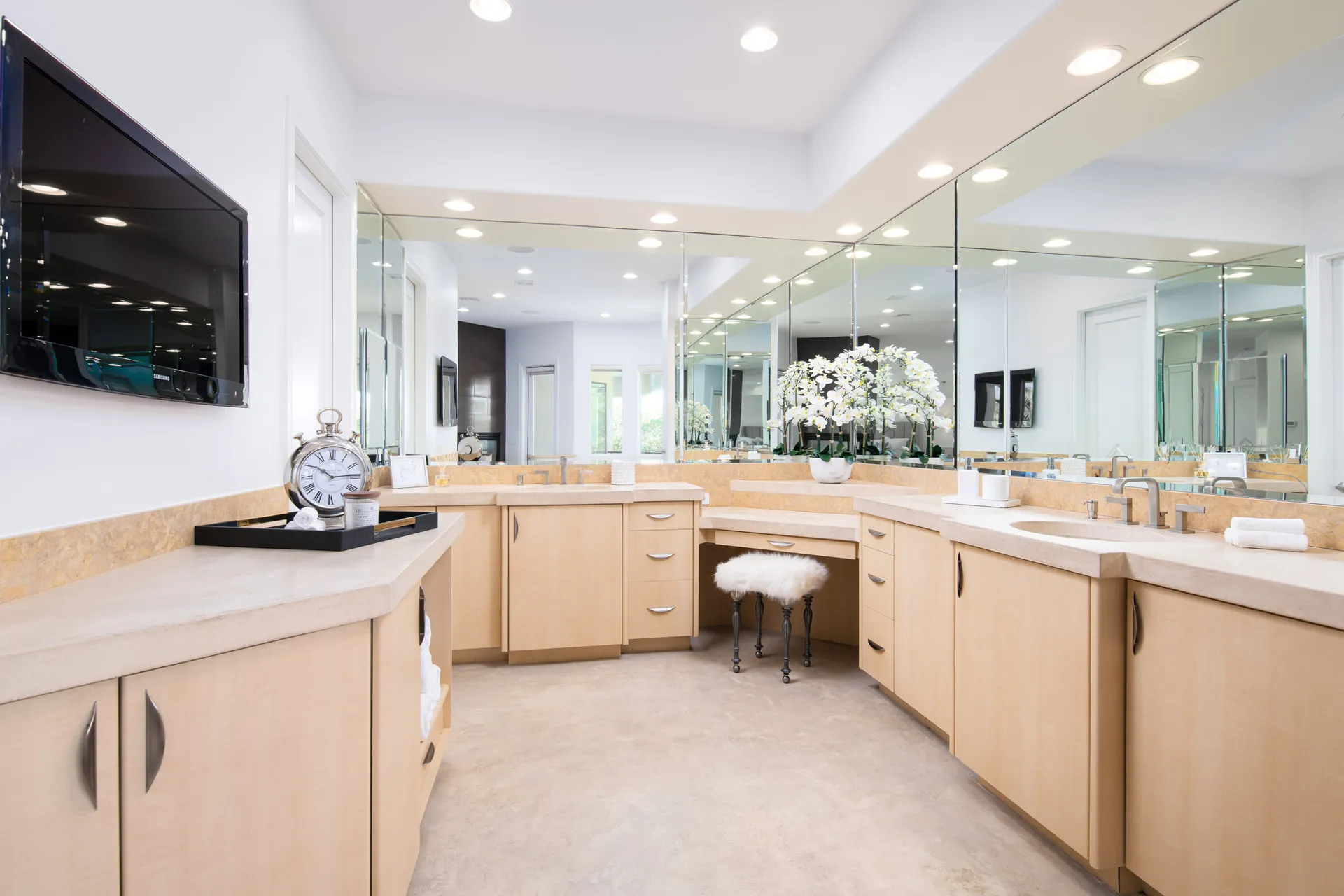 A large master bathroom with light wood cabinetry, a mirrored vanity, a plush fur stool, and decorative accents, including a silver clock and orchids