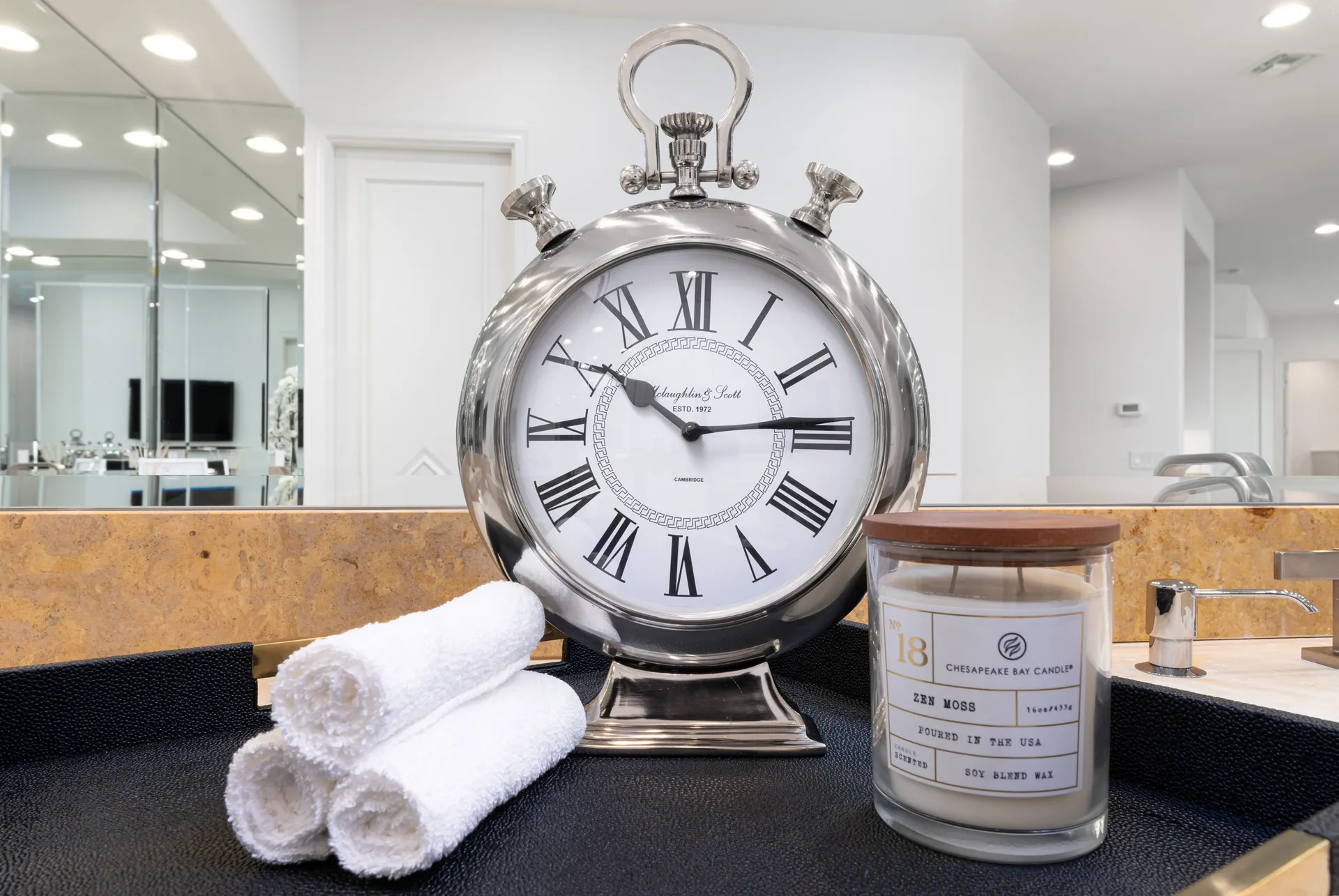 A bathroom vanity styled with a silver vintage clock, a Zen Moss scented candle, and neatly rolled white towels on a black tray.