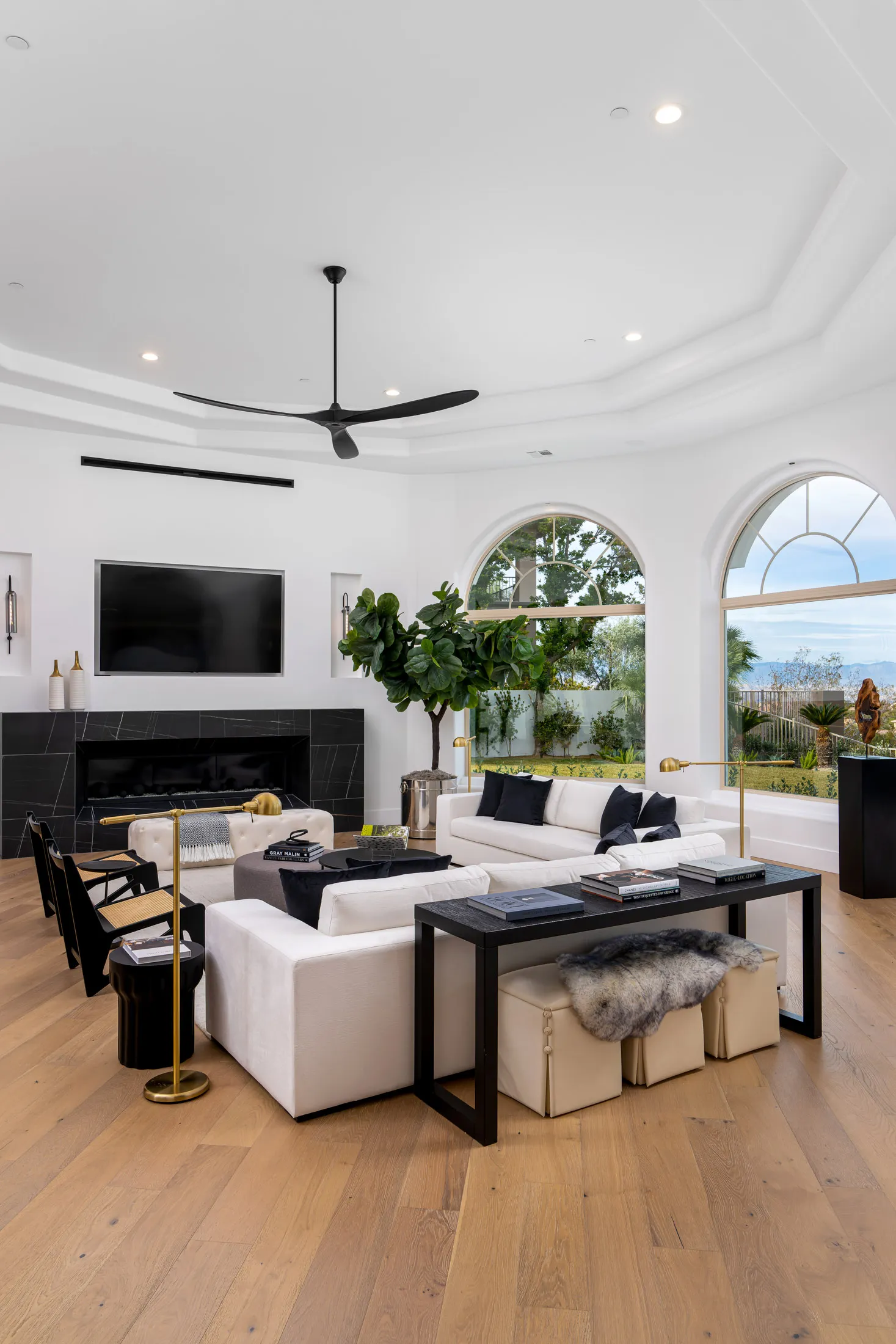 Living room with white sofas, black chairs, a fireplace, and a large potted plant. Large arched windows overlook a scenic view, and a ceiling fan is visible. The room has wooden flooring and a rug, creating a spacious and elegant living space.
