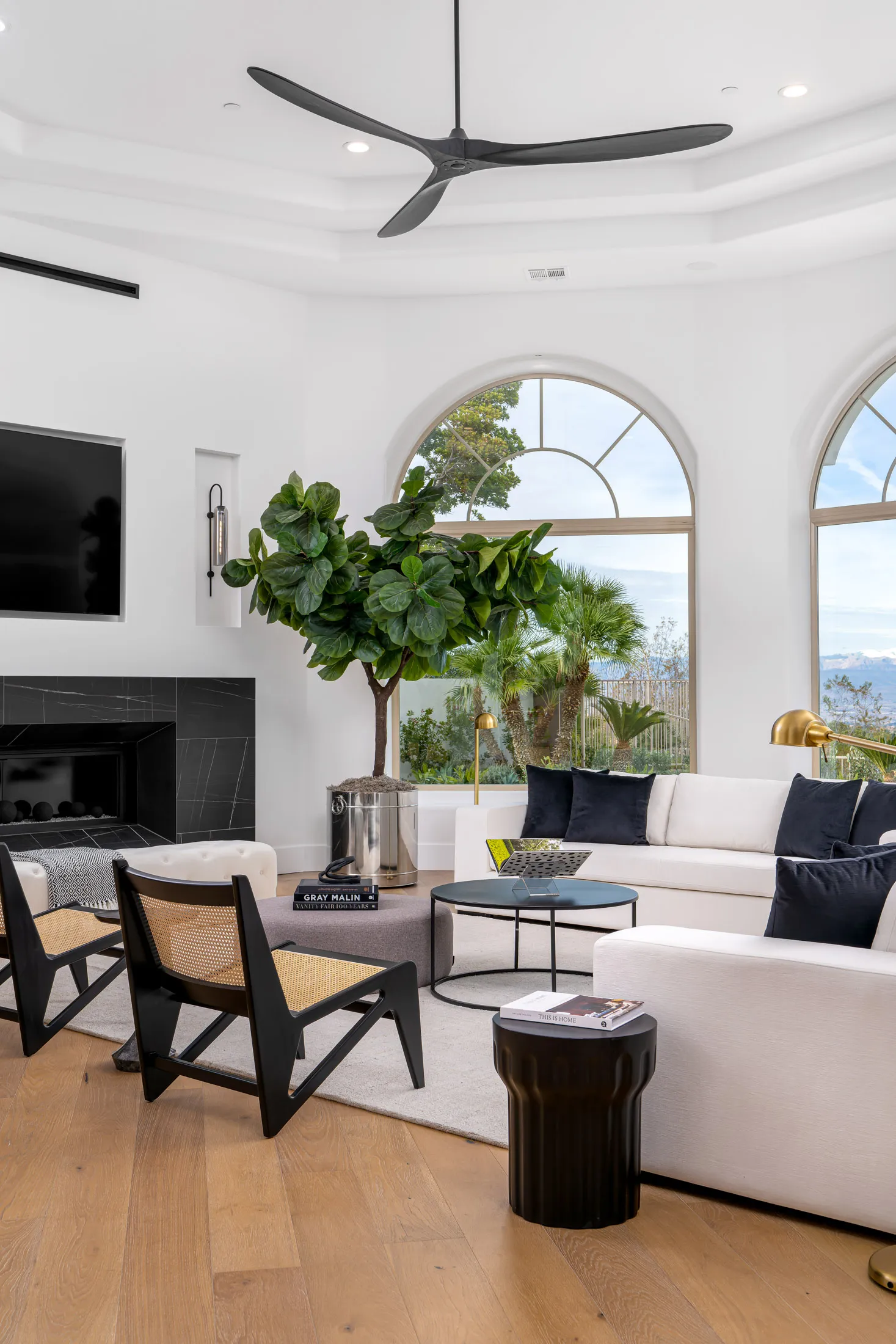 Living room with white sofas, black chairs, a fireplace, and a large potted plant. Large arched windows overlook a scenic view, and a ceiling fan is visible. The room has wooden flooring and a rug, creating a spacious and elegant living space.