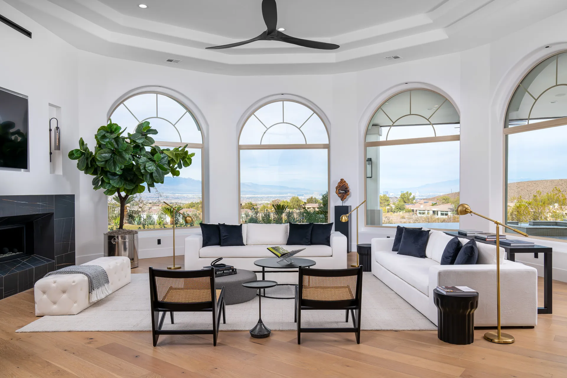 Living room with white sofas, black chairs, a fireplace, and a large potted plant. Large arched windows overlook a scenic view, and a ceiling fan is visible. The room has wooden flooring and a rug, creating a spacious and elegant living space.