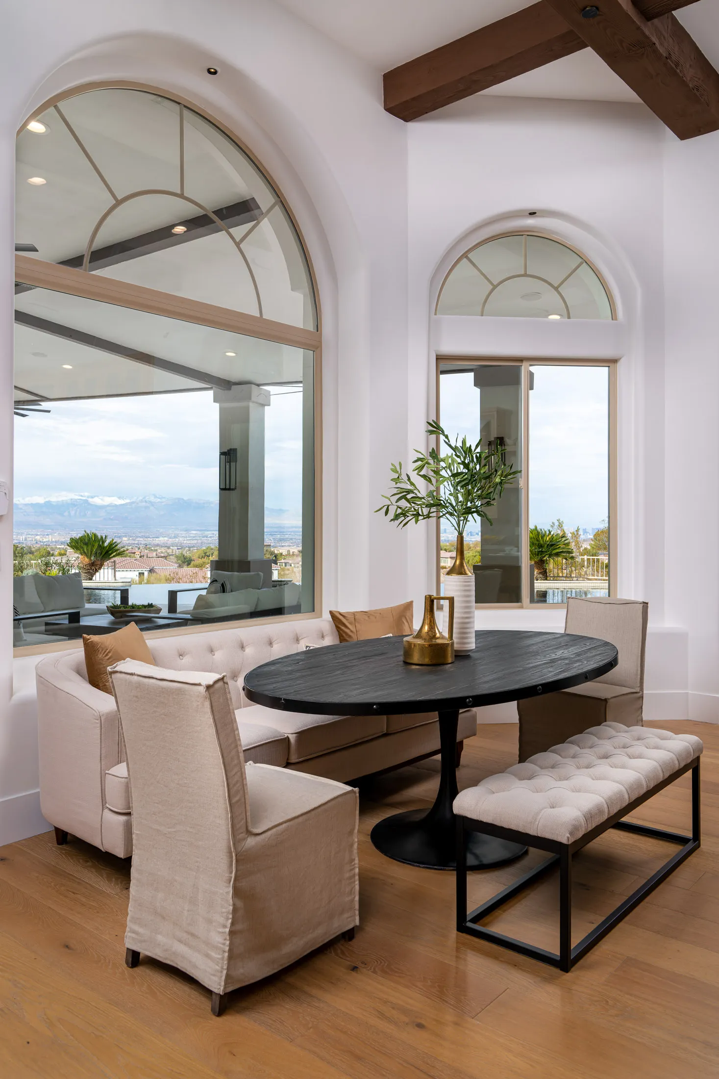 Dining area with a round table, beige chairs, and a large arched window overlooking a scenic view. A potted plant is on the table, and the room features wooden flooring, creating a warm and inviting dining space.