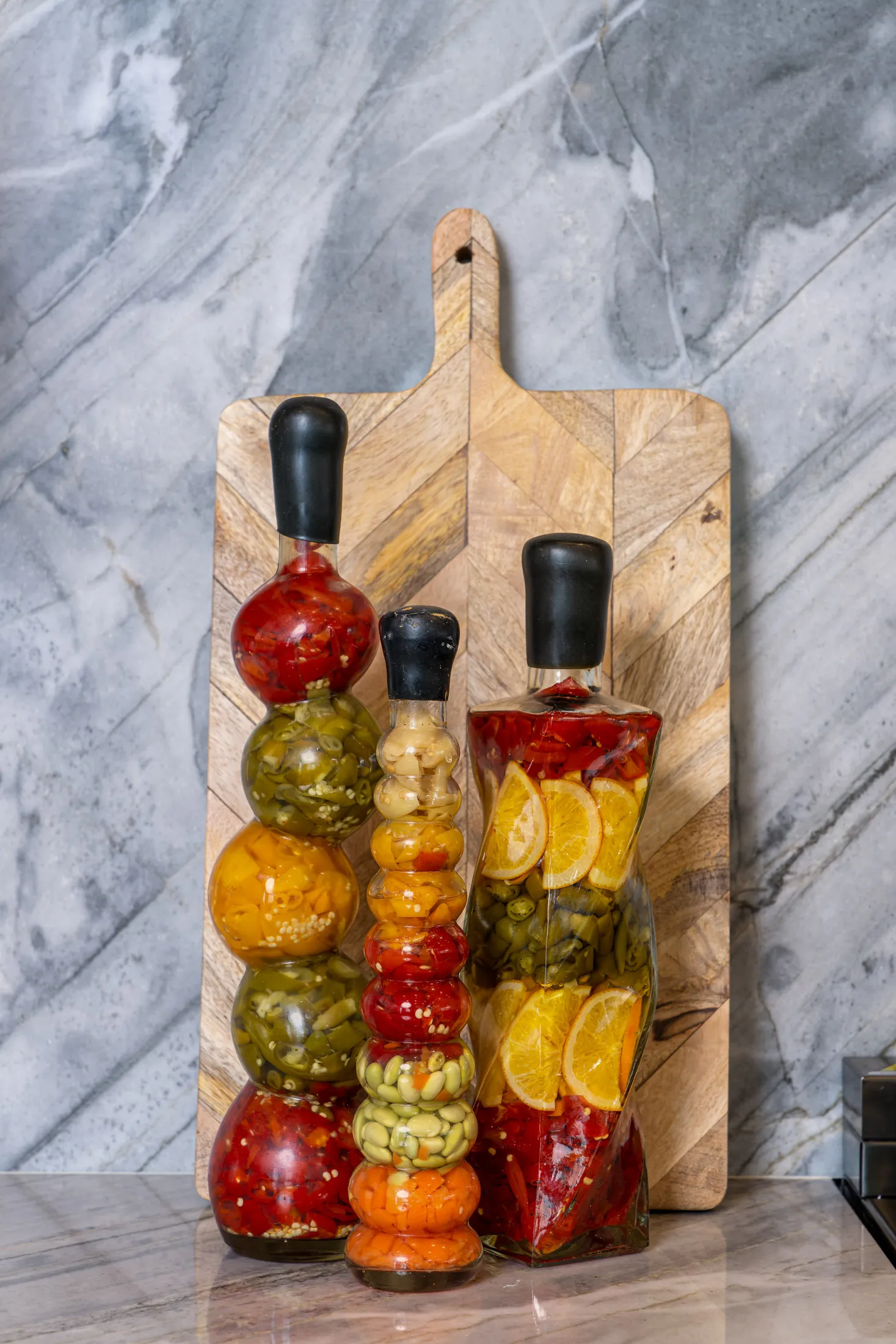 Colorful peppers and citrus slices displayed in decorative glass bottles on a wooden board.