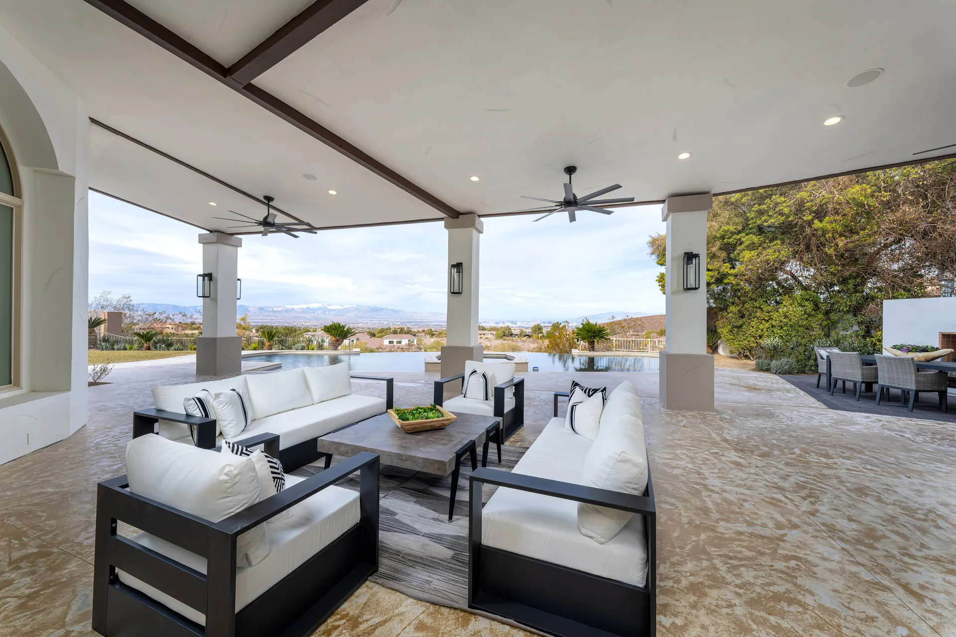 Outdoor seating area with multiple sofas, chairs, and a coffee table under a covered patio. The area overlooks a scenic view of mountains and a landscaped garden, providing a comfortable and inviting outdoor space.