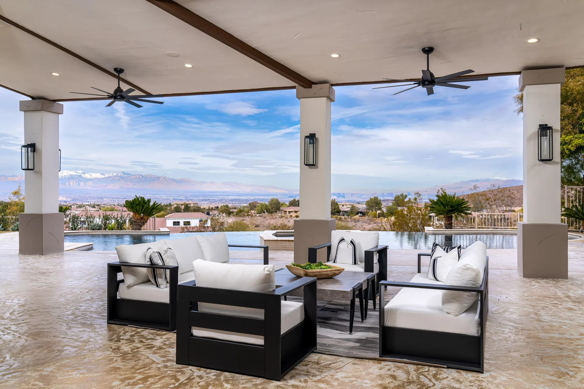A covered outdoor seating area with white cushions and a panoramic desert and mountain view beyond an infinity pool.