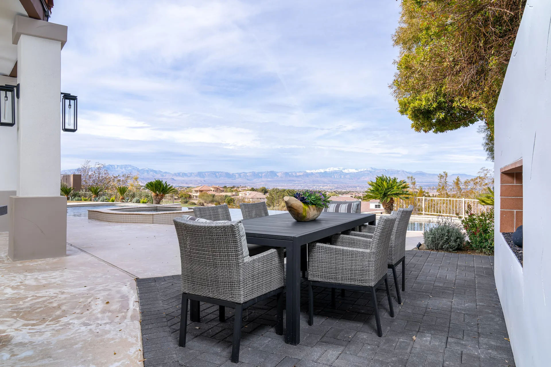 Outdoor patio with a rectangular dining table and six wicker chairs. The patio overlooks a scenic view of mountains and a landscaped garden, offering a serene and inviting outdoor dining space.