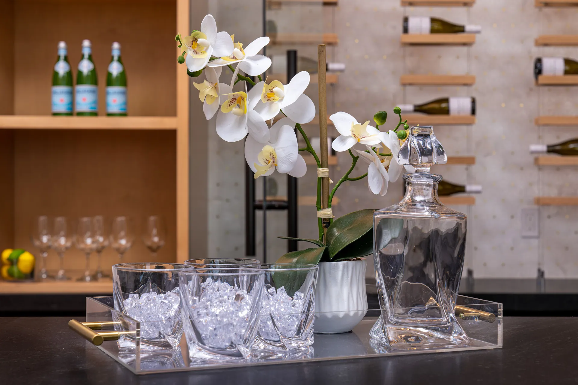 Close-up of a bar setup with a decanter, glasses filled with ice, and a white orchid in a vase. Shelves with bottles and glasses are visible in the background, creating an elegant and inviting bar area.