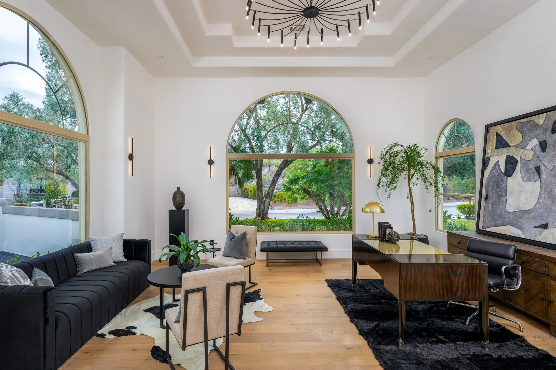 A stylish office with a black sofa, a wooden desk, and large arched windows overlooking lush greenery.