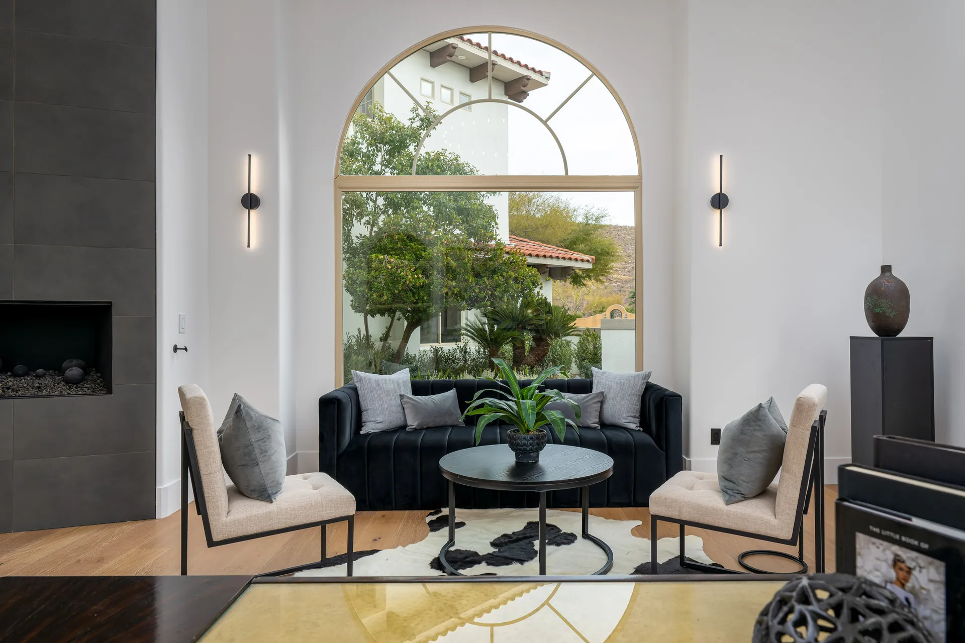 Living room with a large arched window, a black sofa, two beige chairs, and a round coffee table. A fireplace is on the left, and a large plant outside the window adds a natural element to the space.