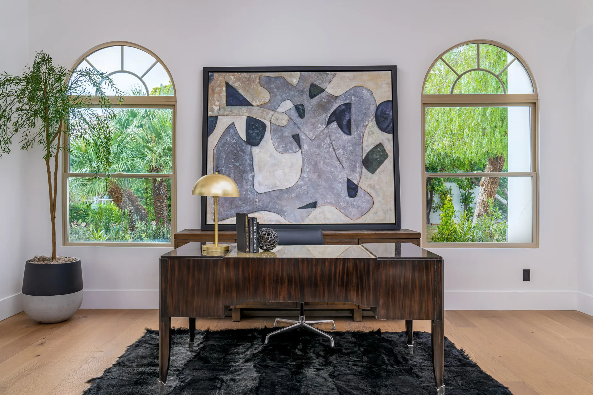 Home office with a glossy wooden desk, a black leather chair, and a black rug. Two large windows with arched frames and a potted plant provide natural light, while a large abstract painting on the wall adds a touch of art.