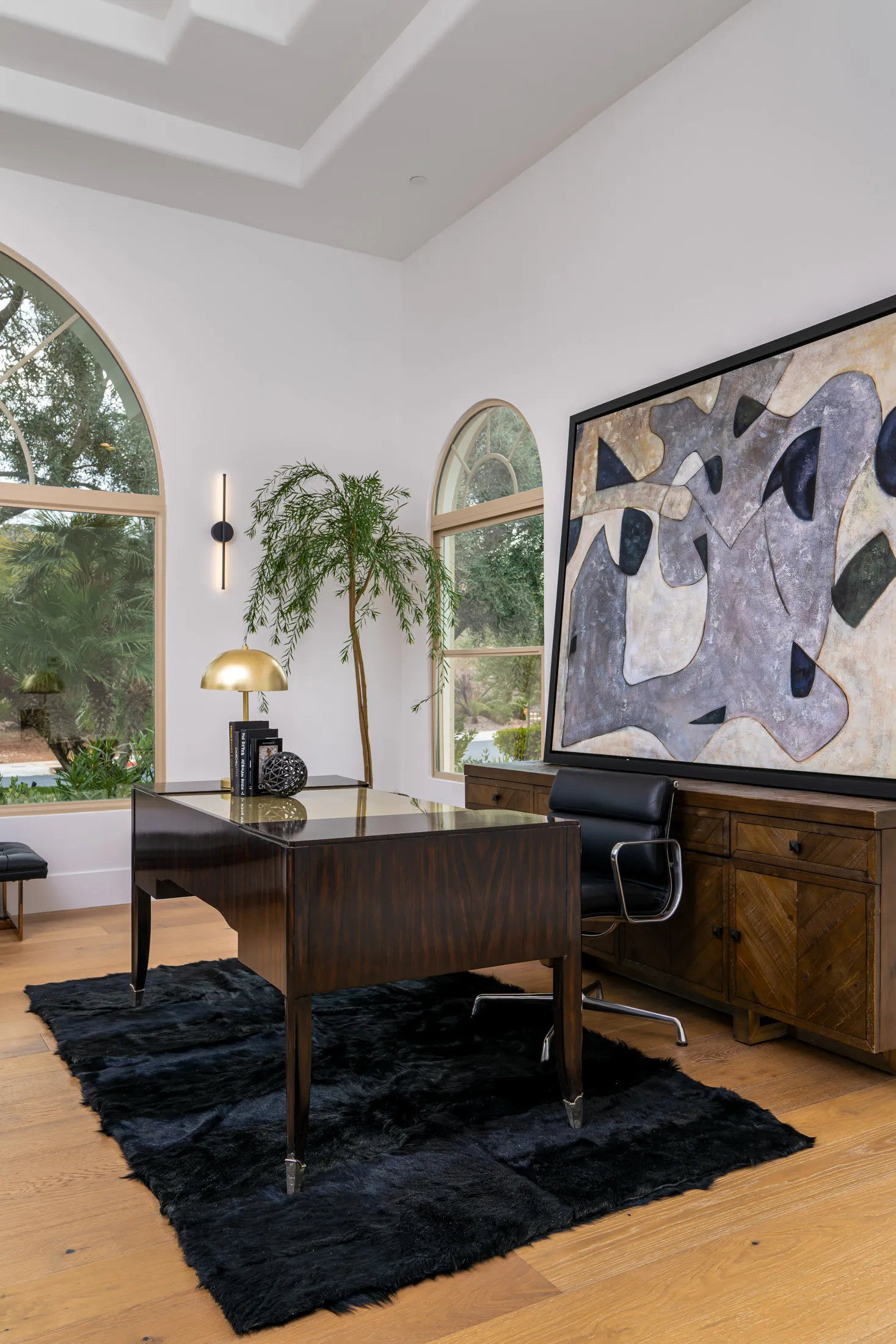 Home office with a glossy wooden desk, a black leather chair, and a black rug. Large windows with arched frames, a potted plant, and a large abstract painting on the wall create a stylish and functional workspace.