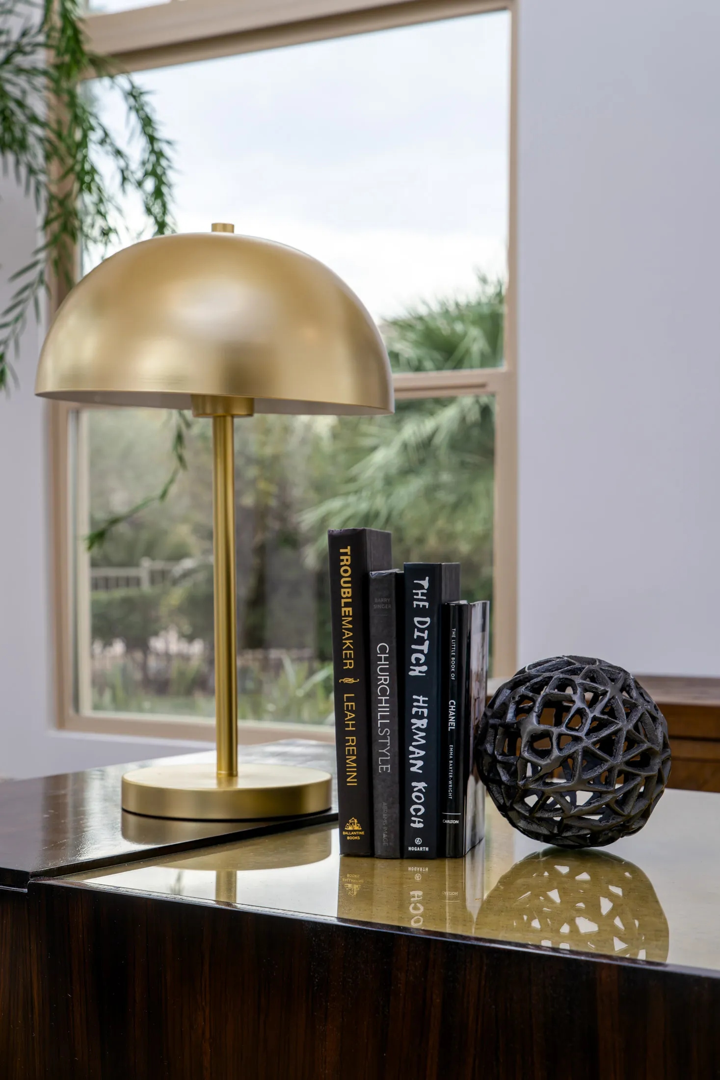 Close-up of a decorative arrangement on a glossy wooden table, featuring a gold lamp, a stack of books, and a decorative sphere. A large window in the background provides natural light, enhancing the elegance of the display.