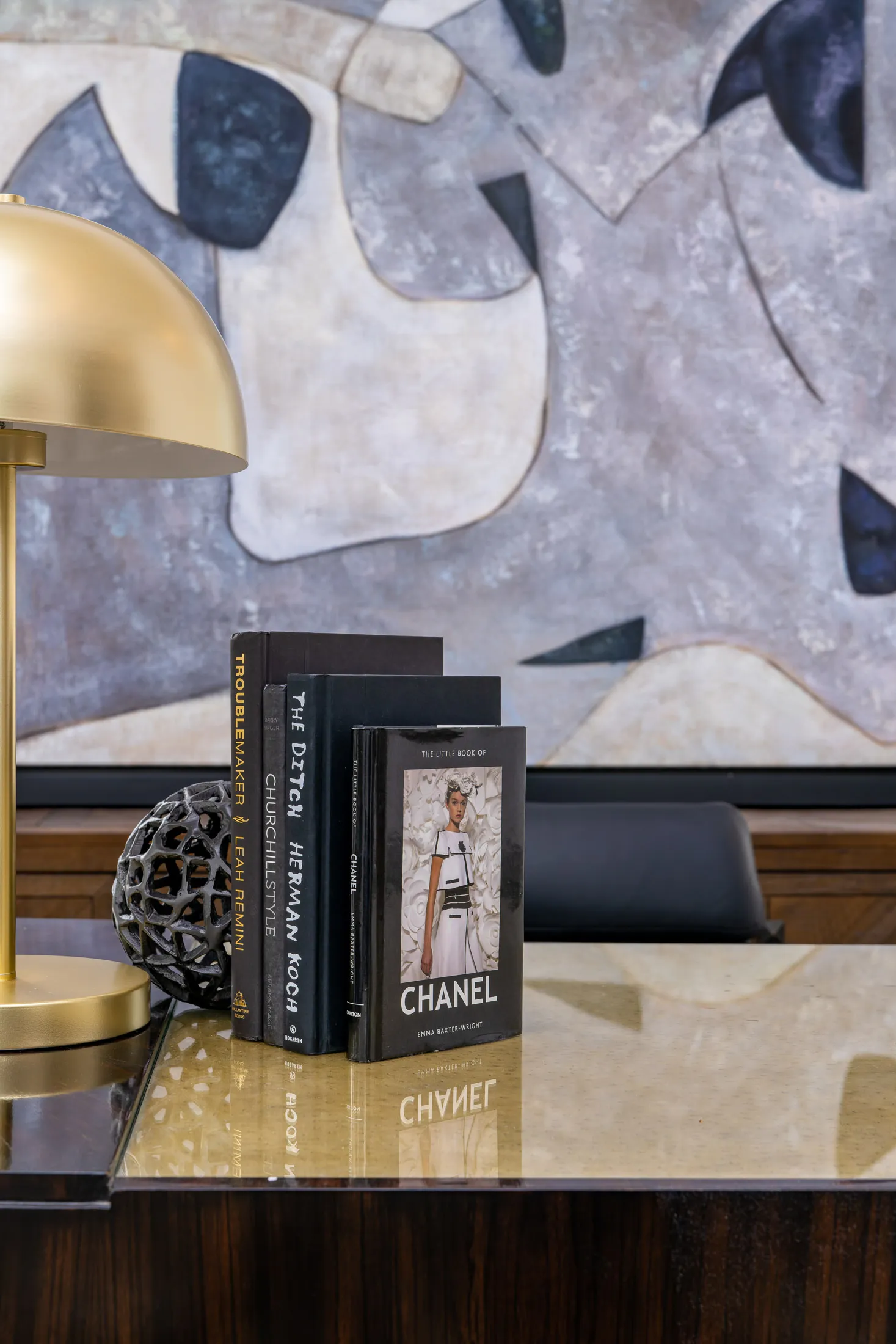 Close-up of a decorative arrangement on a wooden table, featuring a gold lamp, a stack of books with a decorative sphere, and a large abstract painting in the background, creating a sophisticated and artistic display.