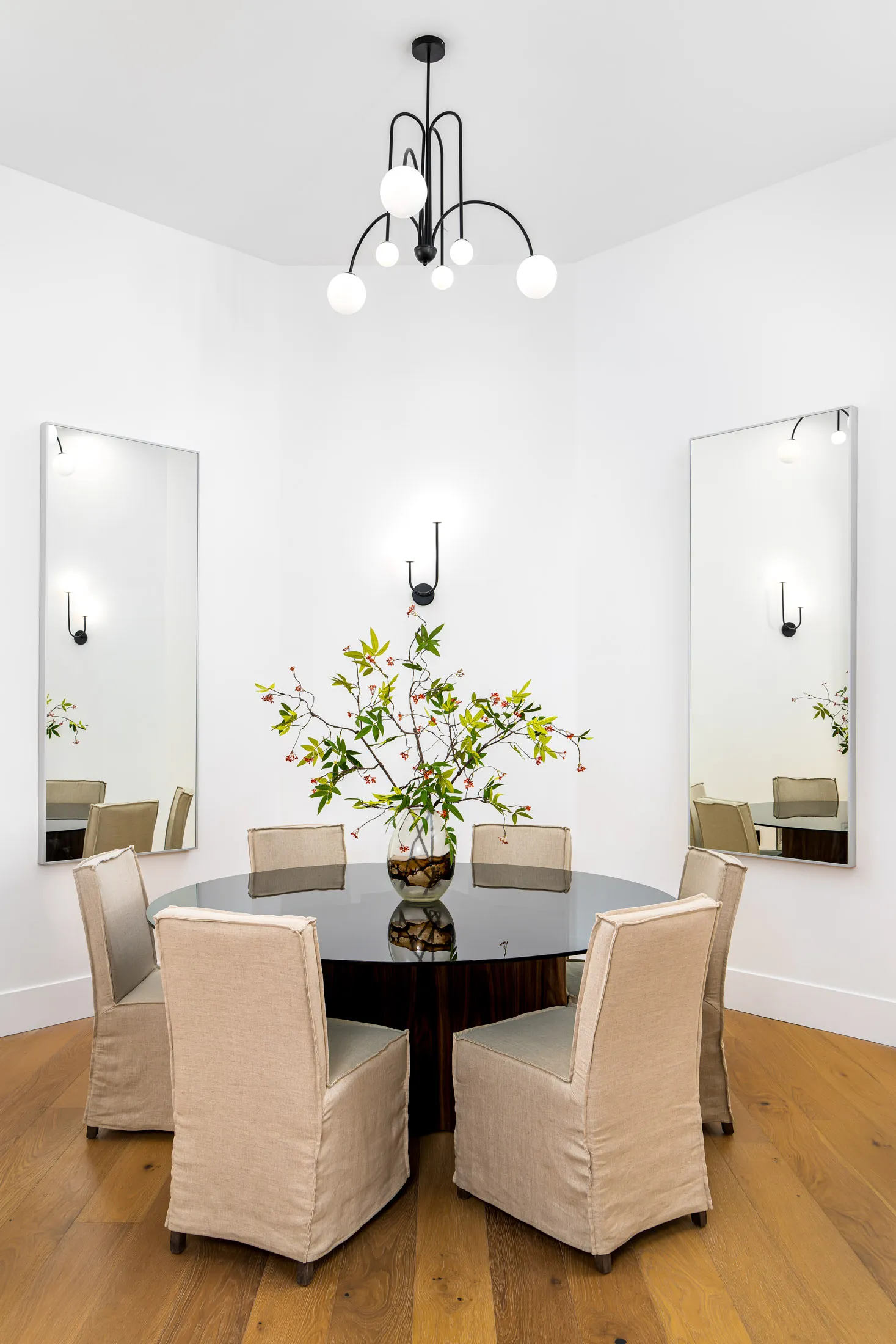 Dining room with a round black table, four beige upholstered chairs, and a vase of flowers in the center. Two large mirrors are on the walls, and a modern chandelier hangs from the ceiling, creating an elegant and inviting space.