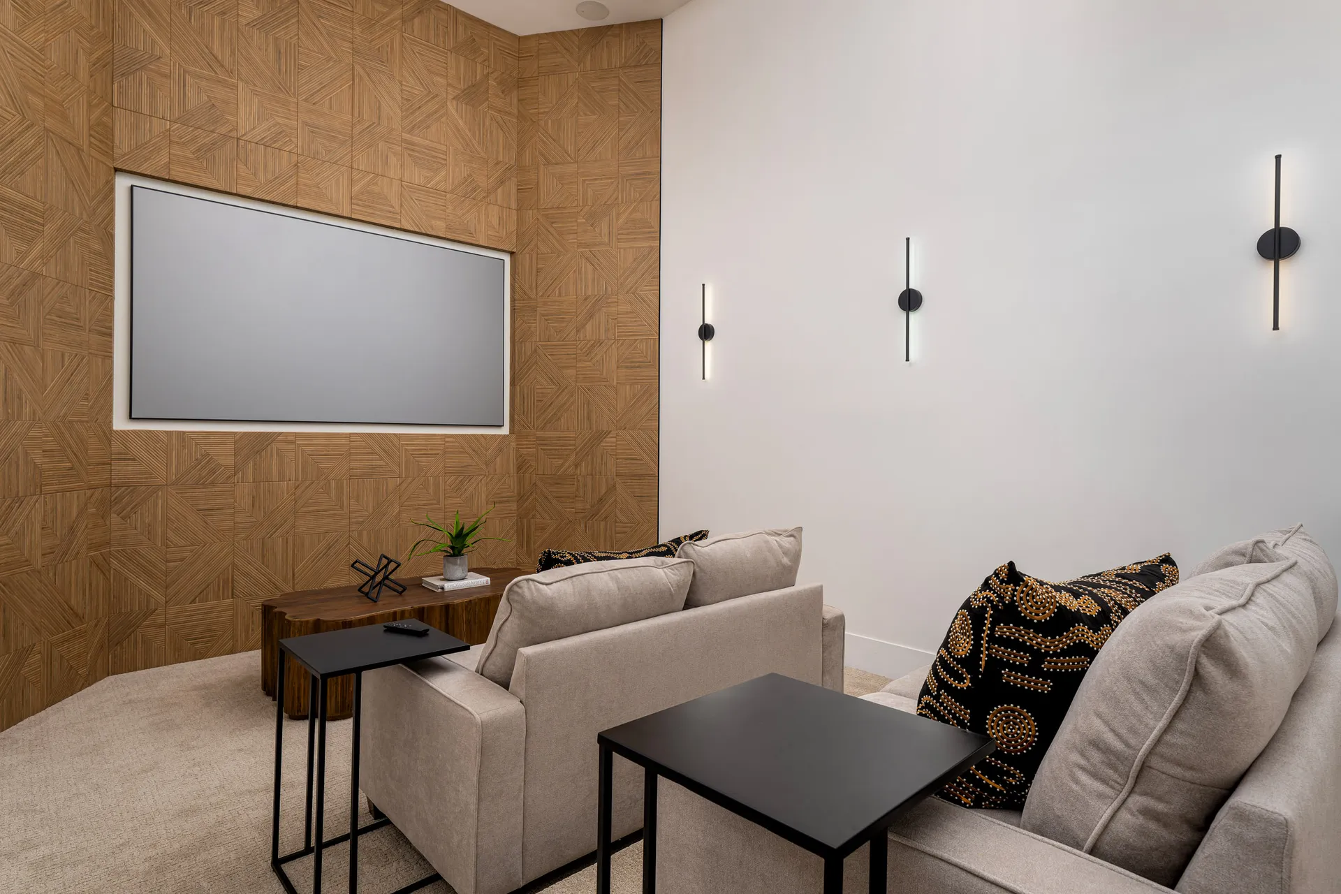 Cozy home theater room with a large screen on the wall, comfortable seating with beige sofas, and a wooden accent wall. The room is illuminated by modern wall sconces, creating a relaxing and inviting atmosphere for entertainment.