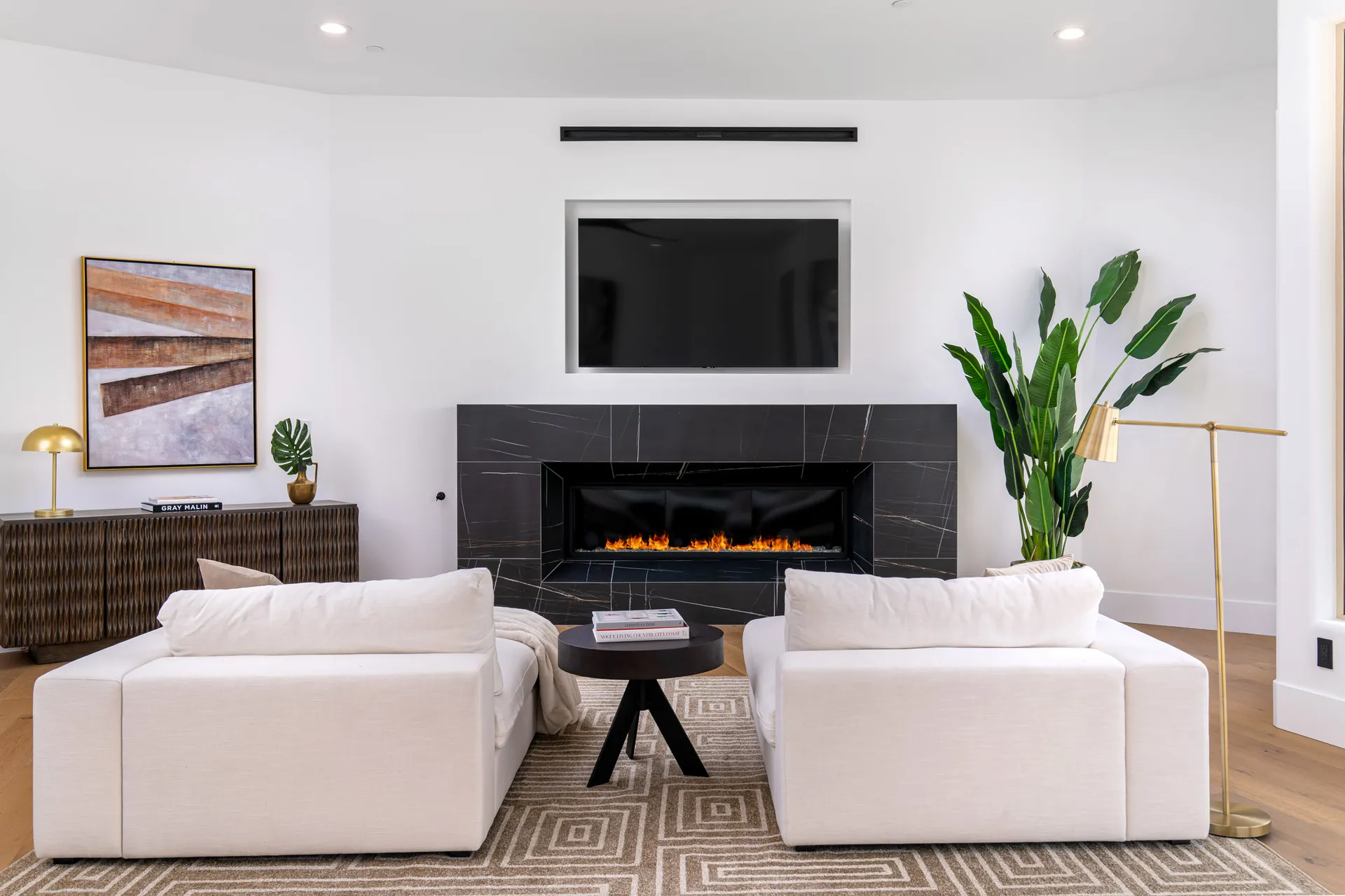 Living room with a modern fireplace, a large TV mounted above it, and two white armchairs facing a black marble coffee table. A large plant is in the corner, and a wooden console table with a lamp and artwork adds to the decor.
