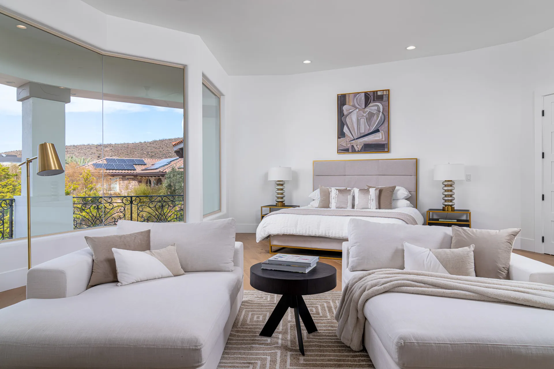 Spacious bedroom with a large bed featuring white linens and beige pillows. Two chaise lounges are positioned at the foot of the bed, accompanied by a circular coffee table. A large window offers a view of the outdoors, enhancing the room's airy ambiance.