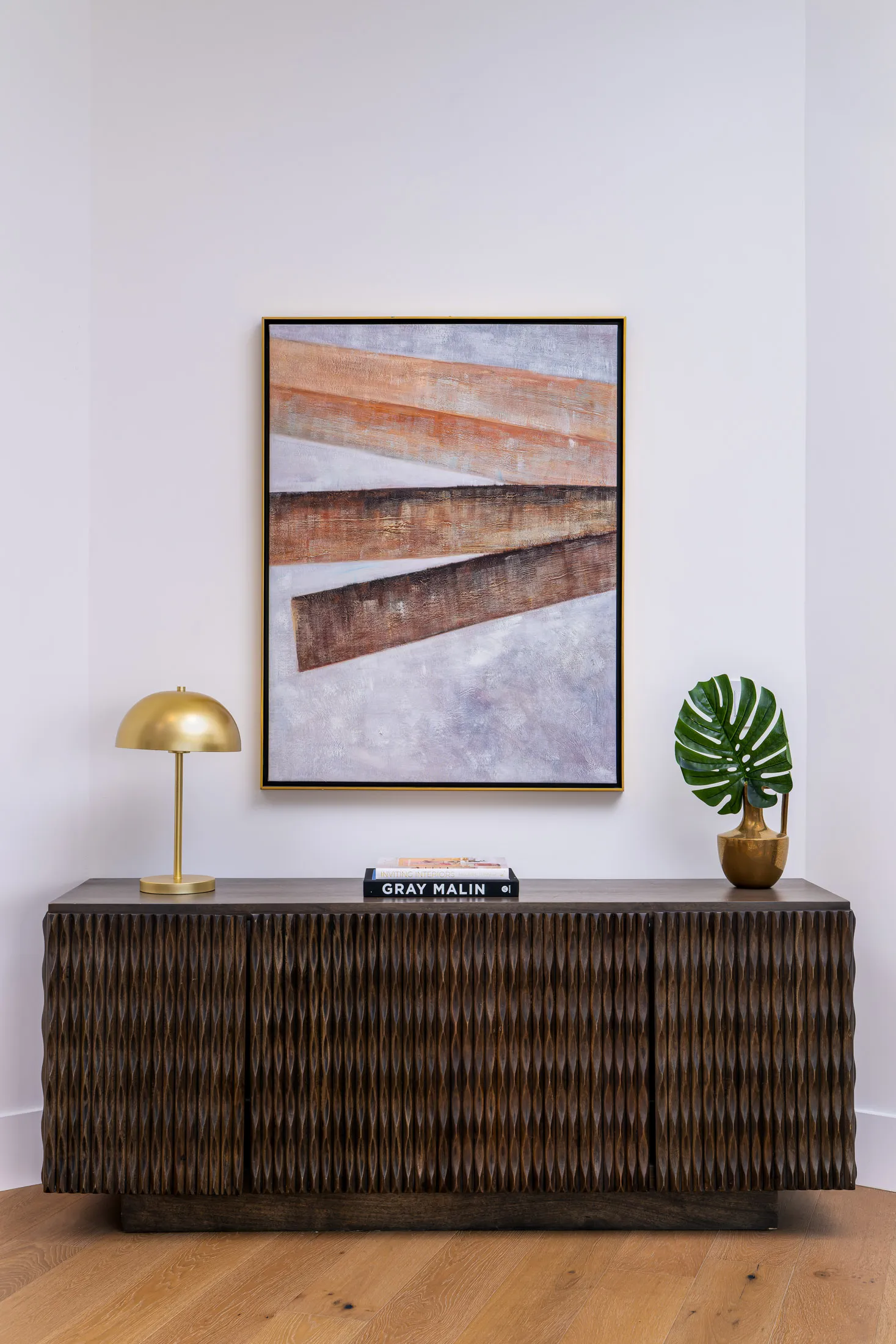 Console table with a textured design, topped with a gold lamp, a potted plant, and a book. Above the table, there is a large abstract painting with earthy tones, creating a stylish and modern display.
