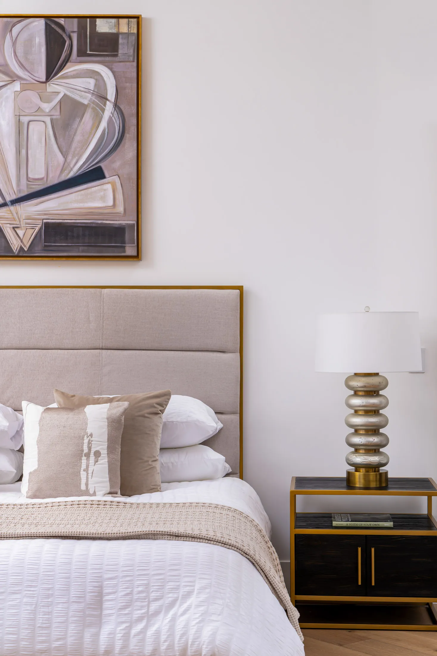 Bedroom with a neatly made bed featuring white linens and beige pillows. A bedside table with a metallic lamp is positioned next to the bed, and a framed abstract artwork hangs on the wall, adding a touch of sophistication.