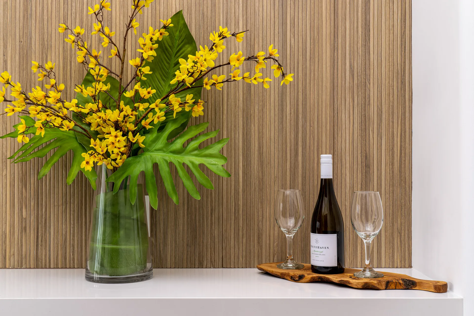 Close-up of a decorative arrangement on a white countertop, featuring a glass vase with yellow flowers and green leaves, accompanied by a wooden tray with a wine bottle and two glasses.