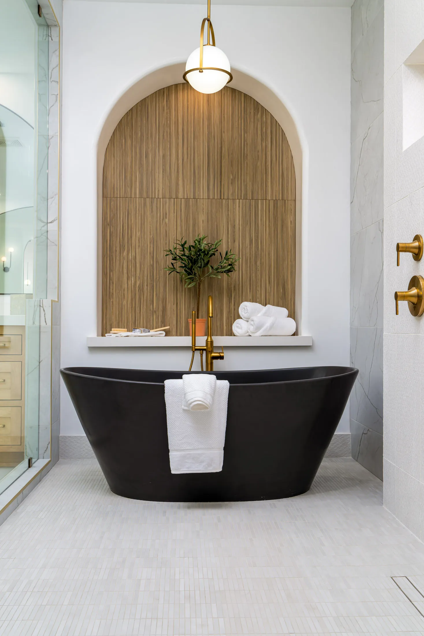 Master bathroom featuring a freestanding black bathtub under an arched niche with a wooden accent wall. A gold pendant light hangs above, and brass fixtures complement the design, creating a luxurious and elegant atmosphere.