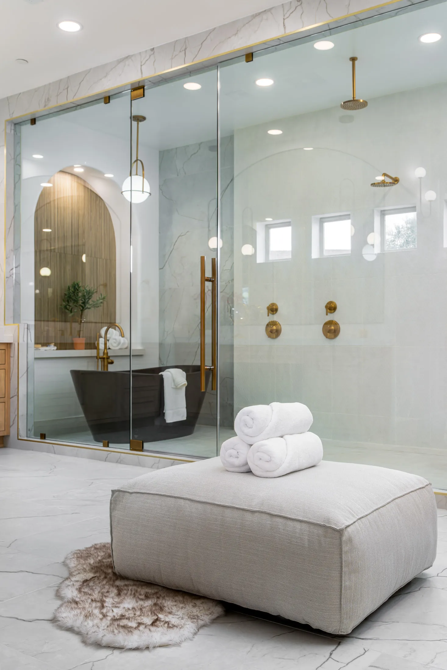 Luxurious bathroom featuring a glass-enclosed shower with brass fixtures, a freestanding bathtub, and a plush ottoman with rolled towels. The walls are made of marble, and there's a fur rug on the floor, adding to the opulent ambiance.