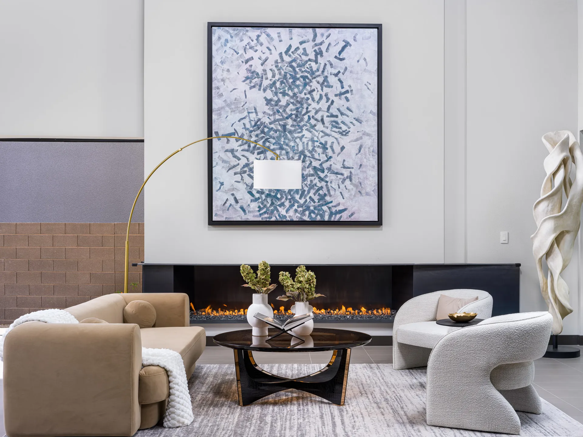 A contemporary living area with a curved tan sofa, a sculptural accent chair, and a long fireplace under a large abstract painting.