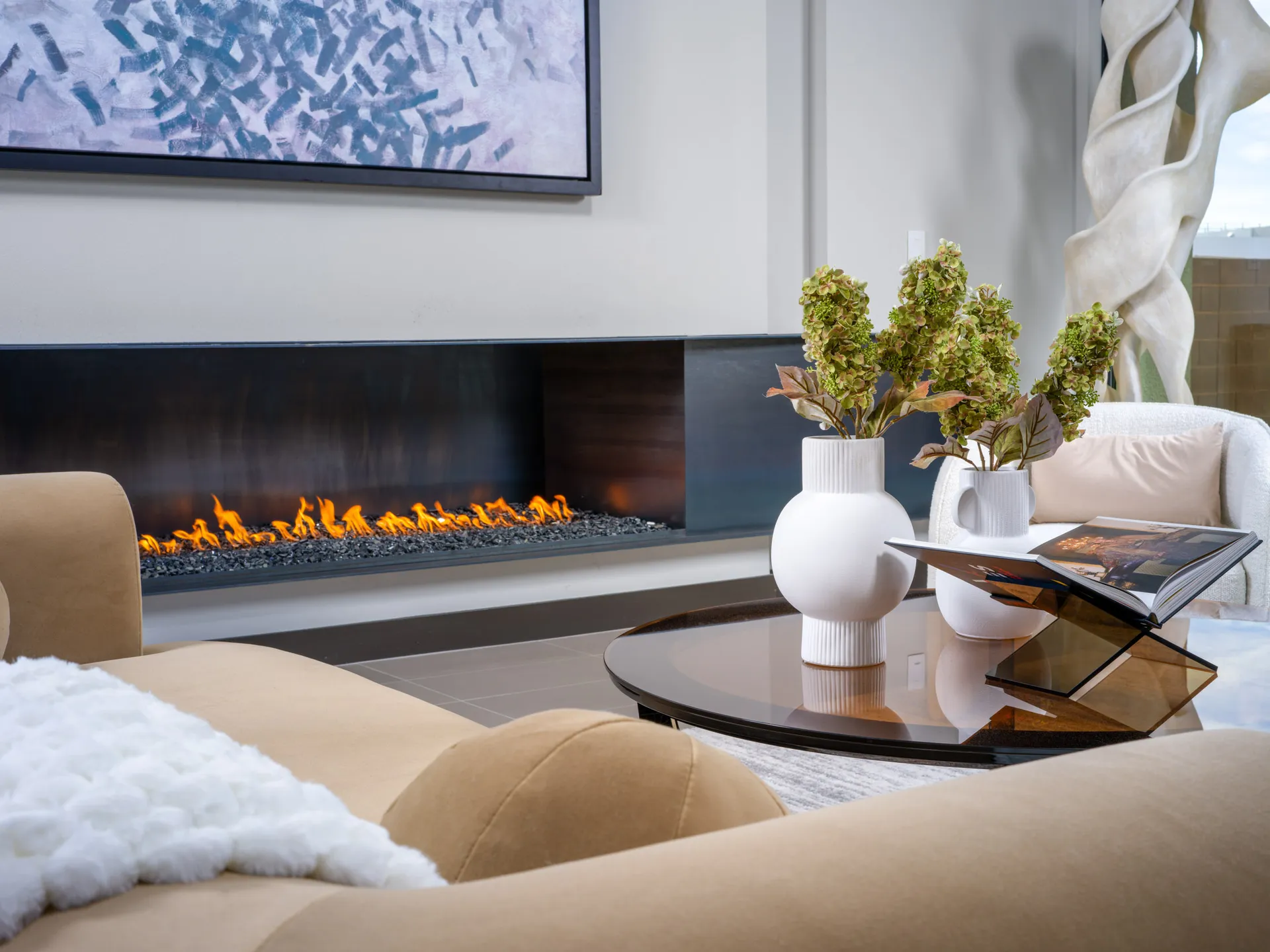 A tan sofa near a long linear fireplace, with a round coffee table displaying decorative vases in the foreground.