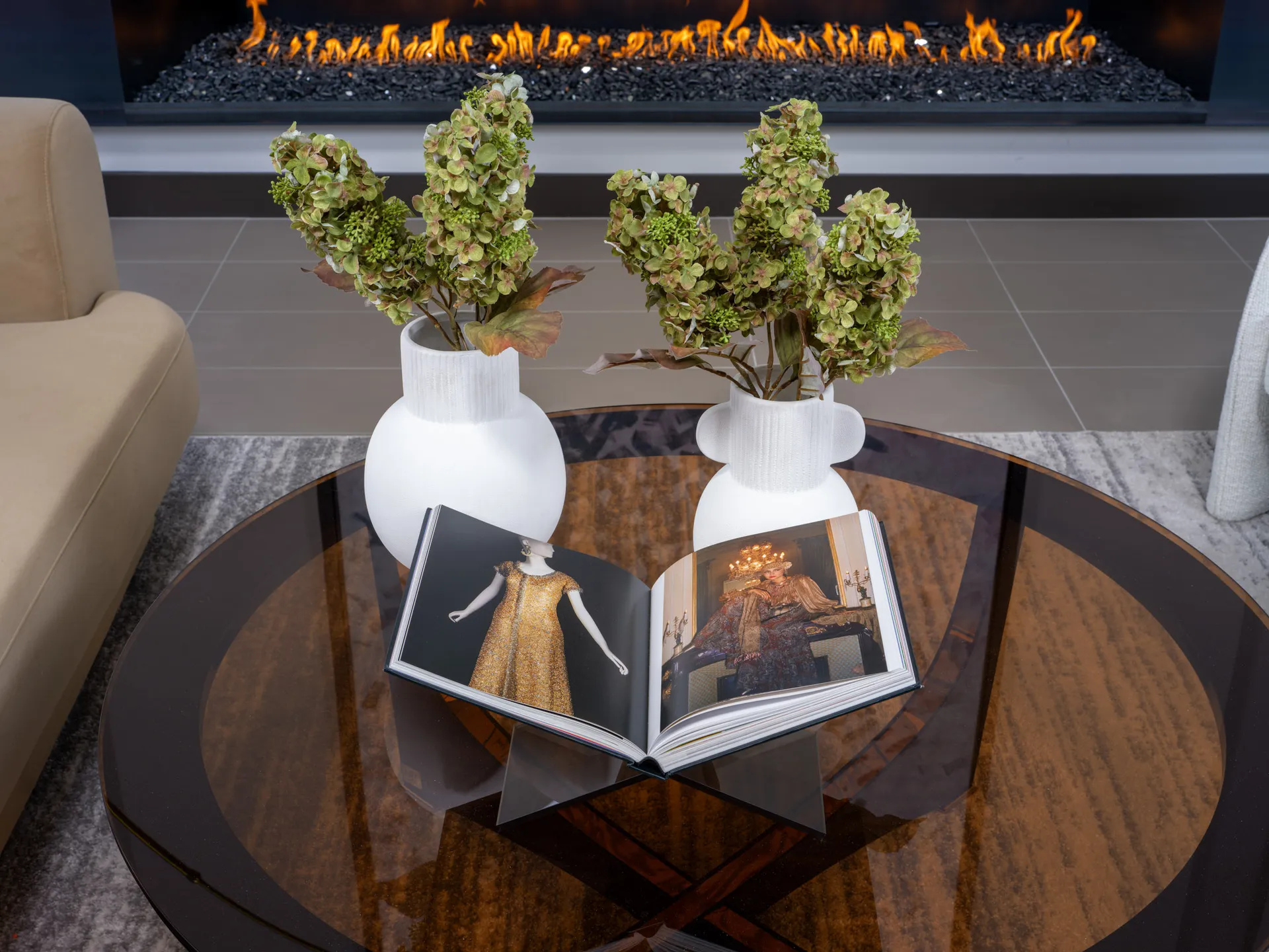 A round coffee table displaying white vases of greenery and an open design book, set near a linear fireplace.