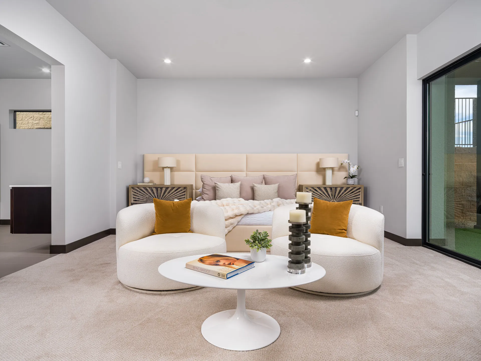 A neutral-toned bedroom with a padded headboard, two curved chairs, and a white coffee table near sliding glass doors.