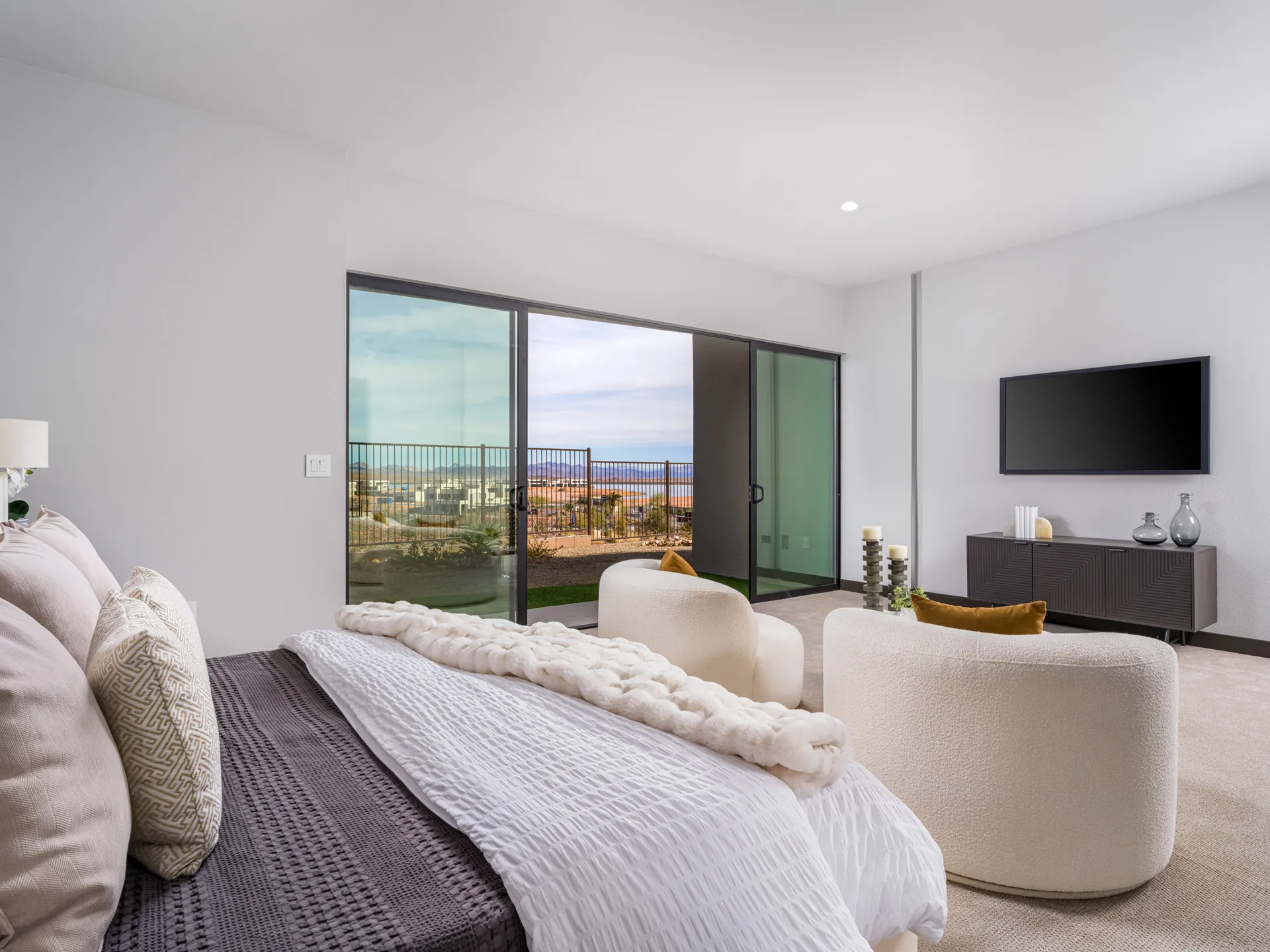 A serene bedroom with a padded headboard and two white chairs facing large sliding doors that reveal desert vistas.