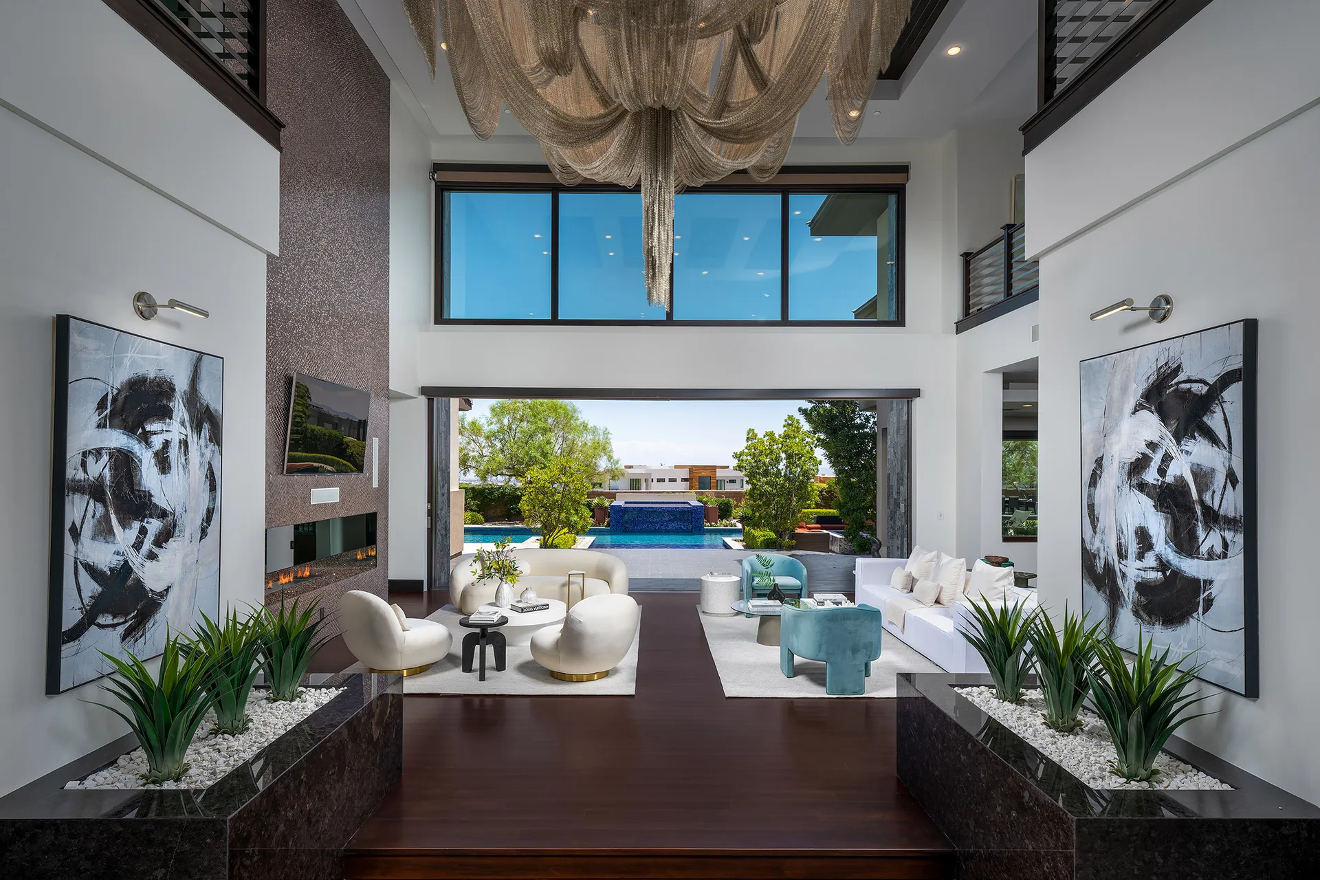 A double-height living room featuring a cascading chandelier, abstract artwork, and plush seating with a view of the pool.