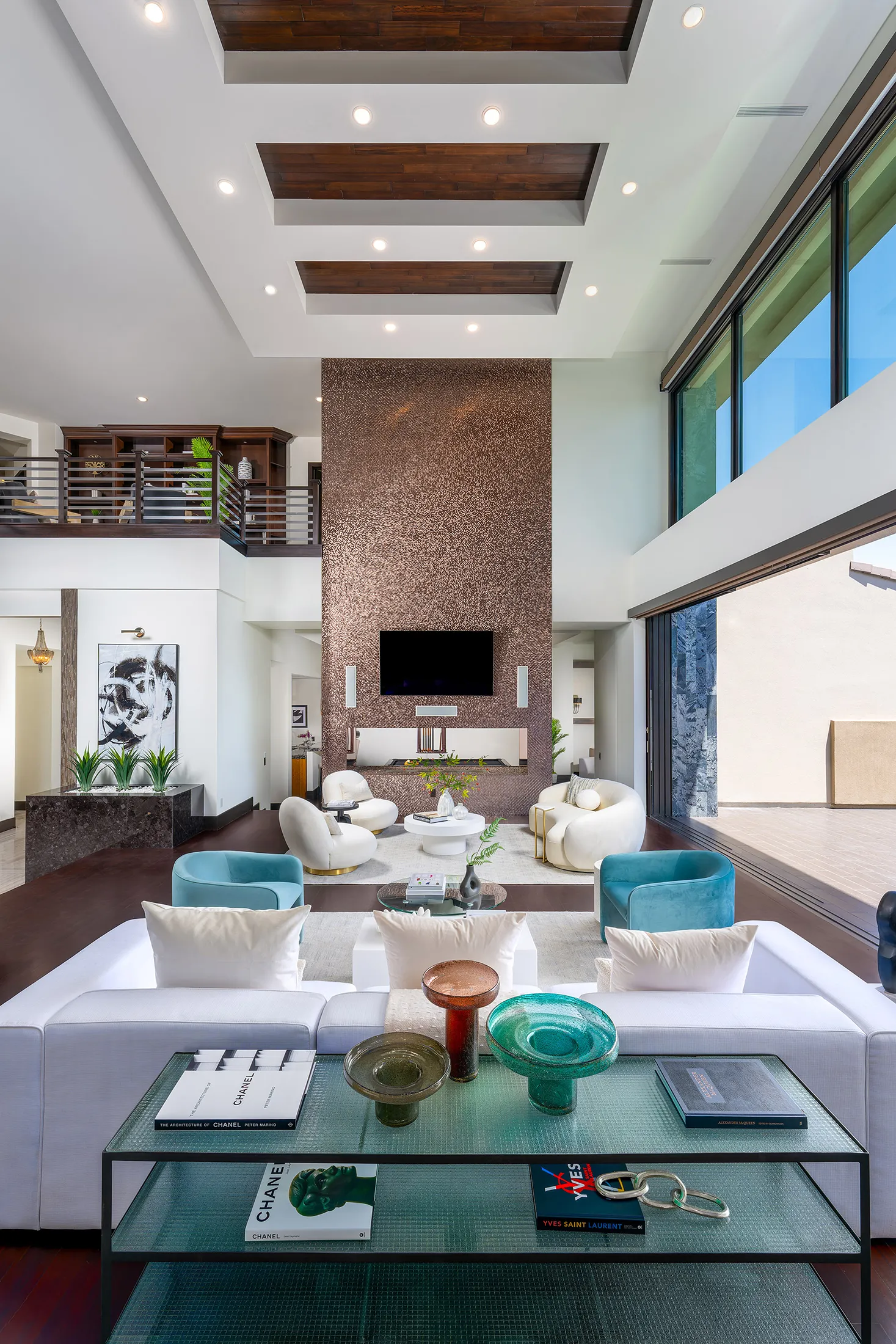 A high-ceiling living area featuring a copper-toned accent wall with a built-in fireplace, plush white seating, and teal chairs below a loft.