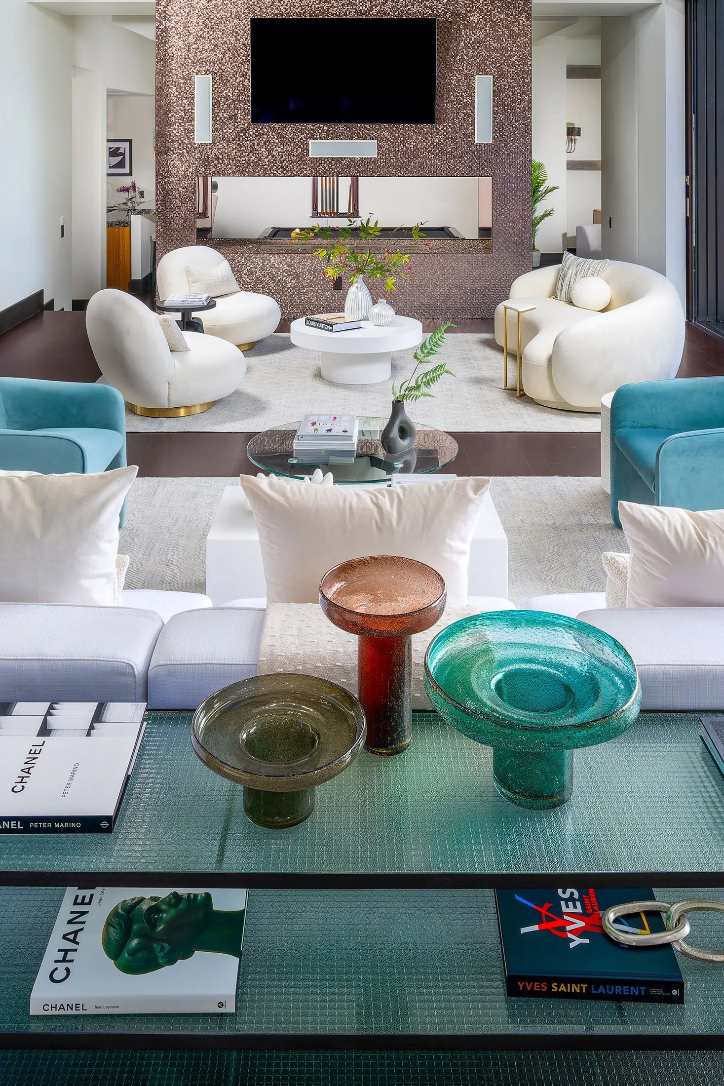 A living room space highlighting a copper-toned fireplace, plush white chairs, and a glass table in the foreground with colorful glass bowls.