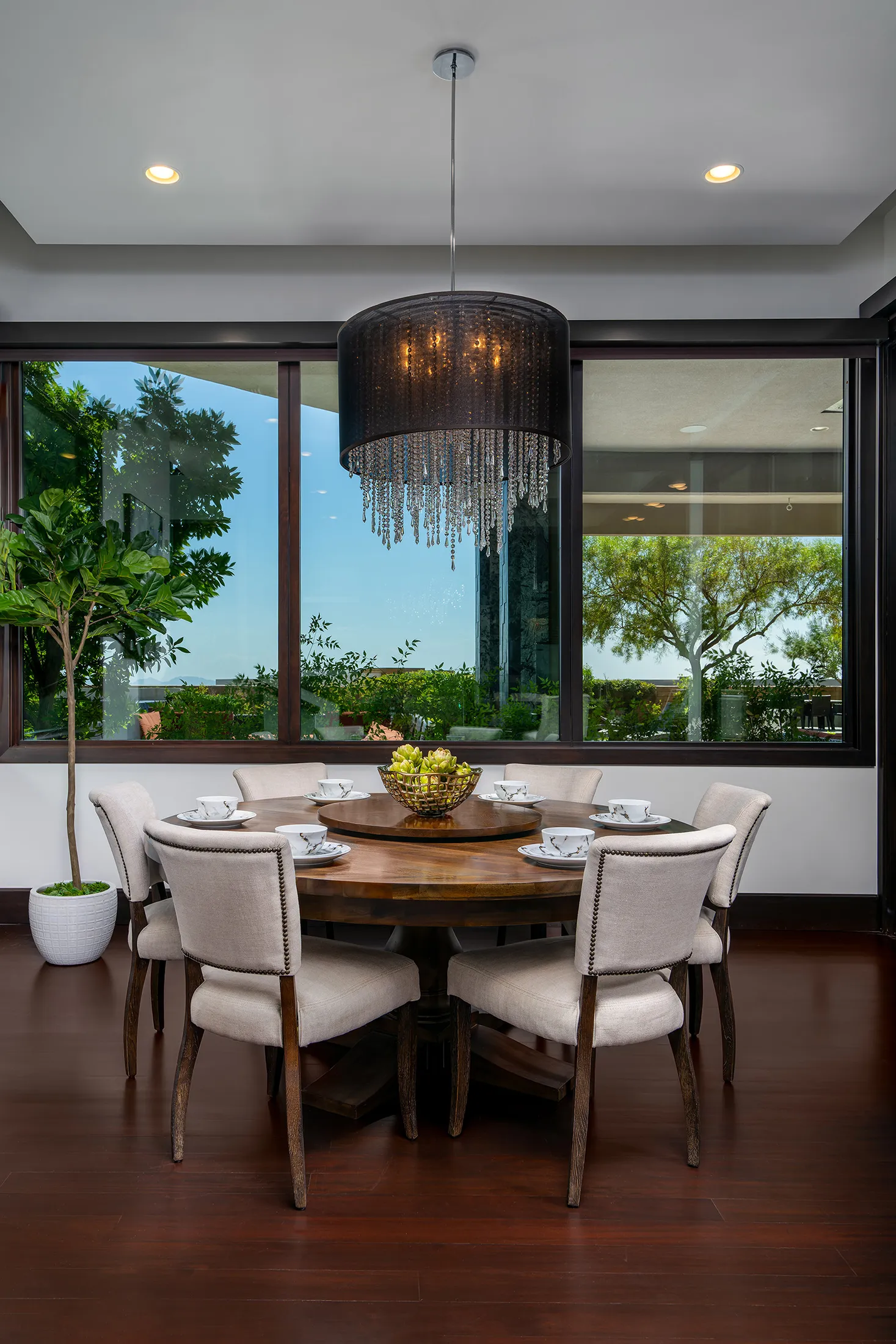 A round wooden dining table with upholstered chairs, a dramatic black chandelier above, and large windows overlooking greenery.